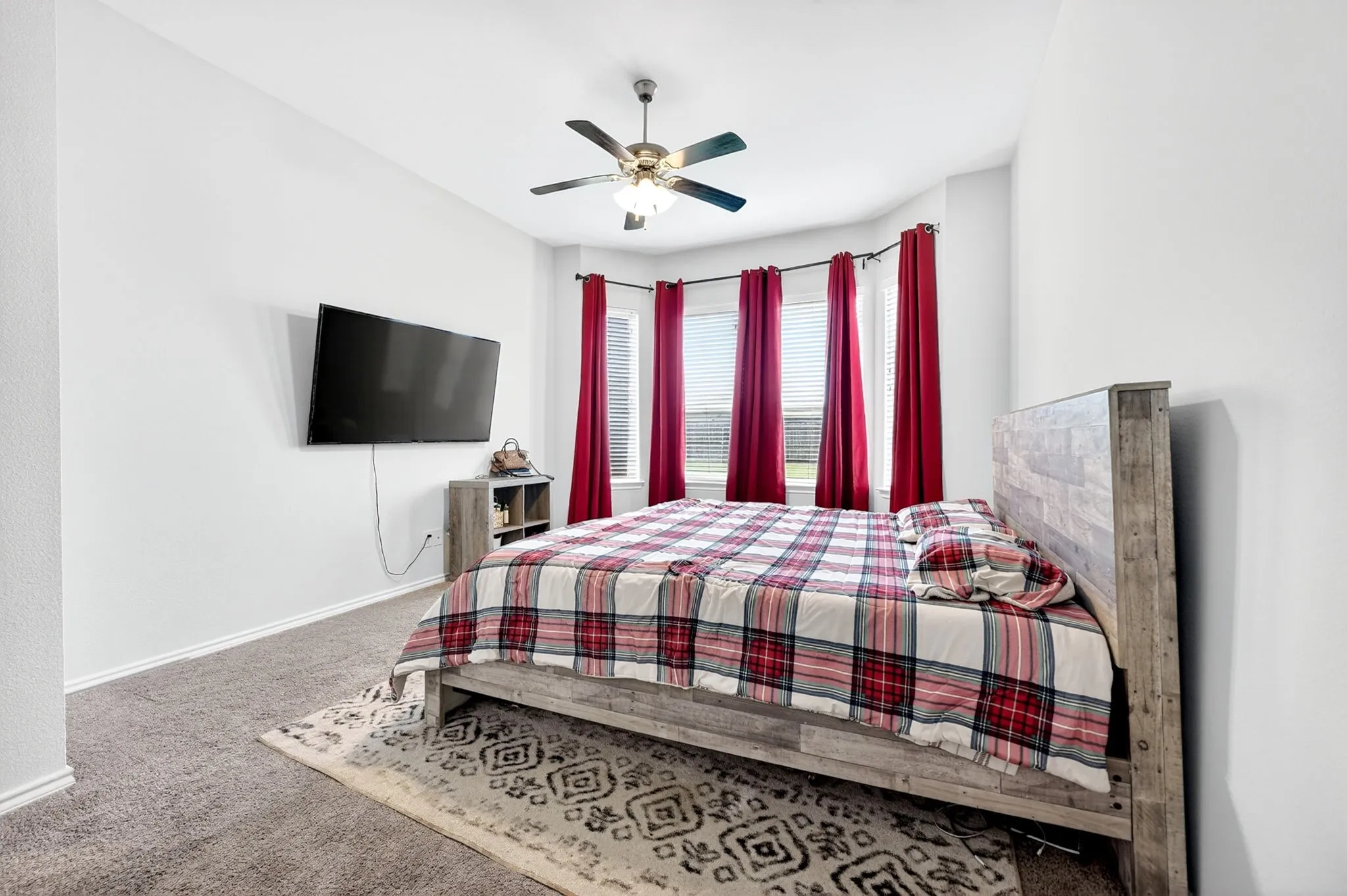 Spacious bedroom featuring light gray walls, plush carpet flooring, a ceiling fan with integrated lighting, and a bay window with multiple vertical blinds