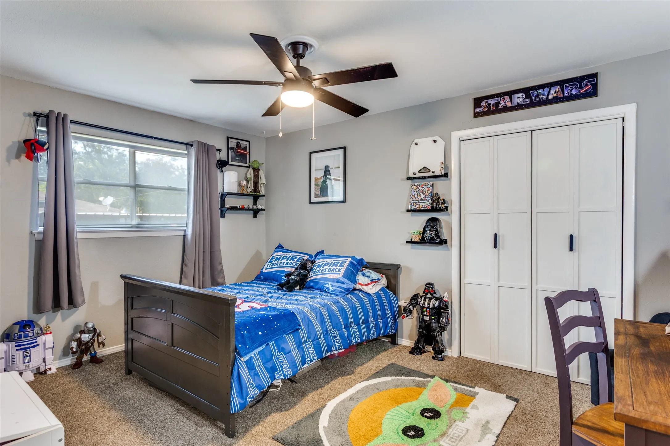 Carpeted bedroom featuring a ceiling fan with light fixture, a window with gray curtains, gray walls, built-in shelving, and bifold closet doors with black hardware