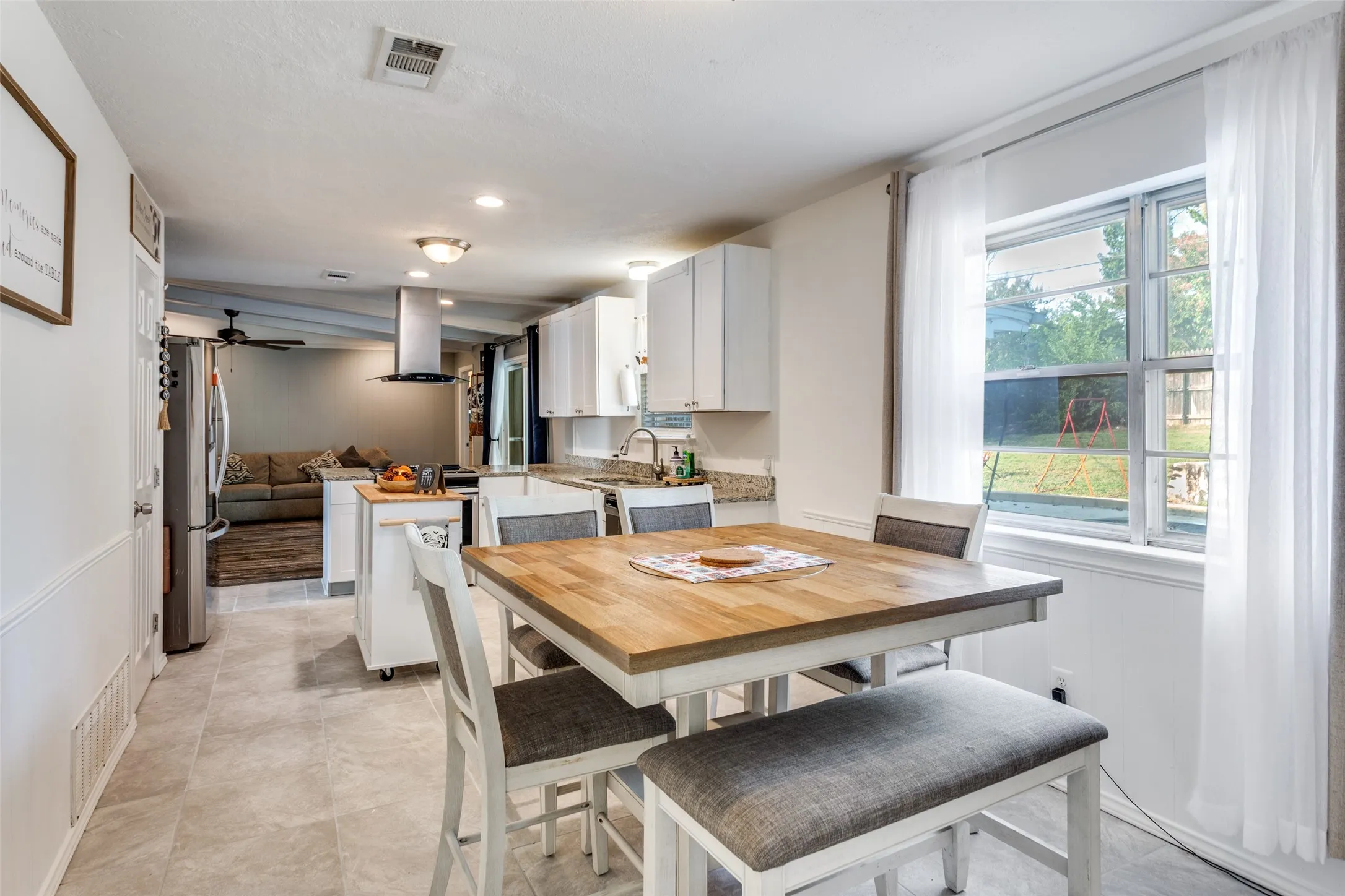 Open-concept kitchen and dining area featuring light-toned tile flooring, white cabinetry, a kitchen island, and a window with sheer white curtains