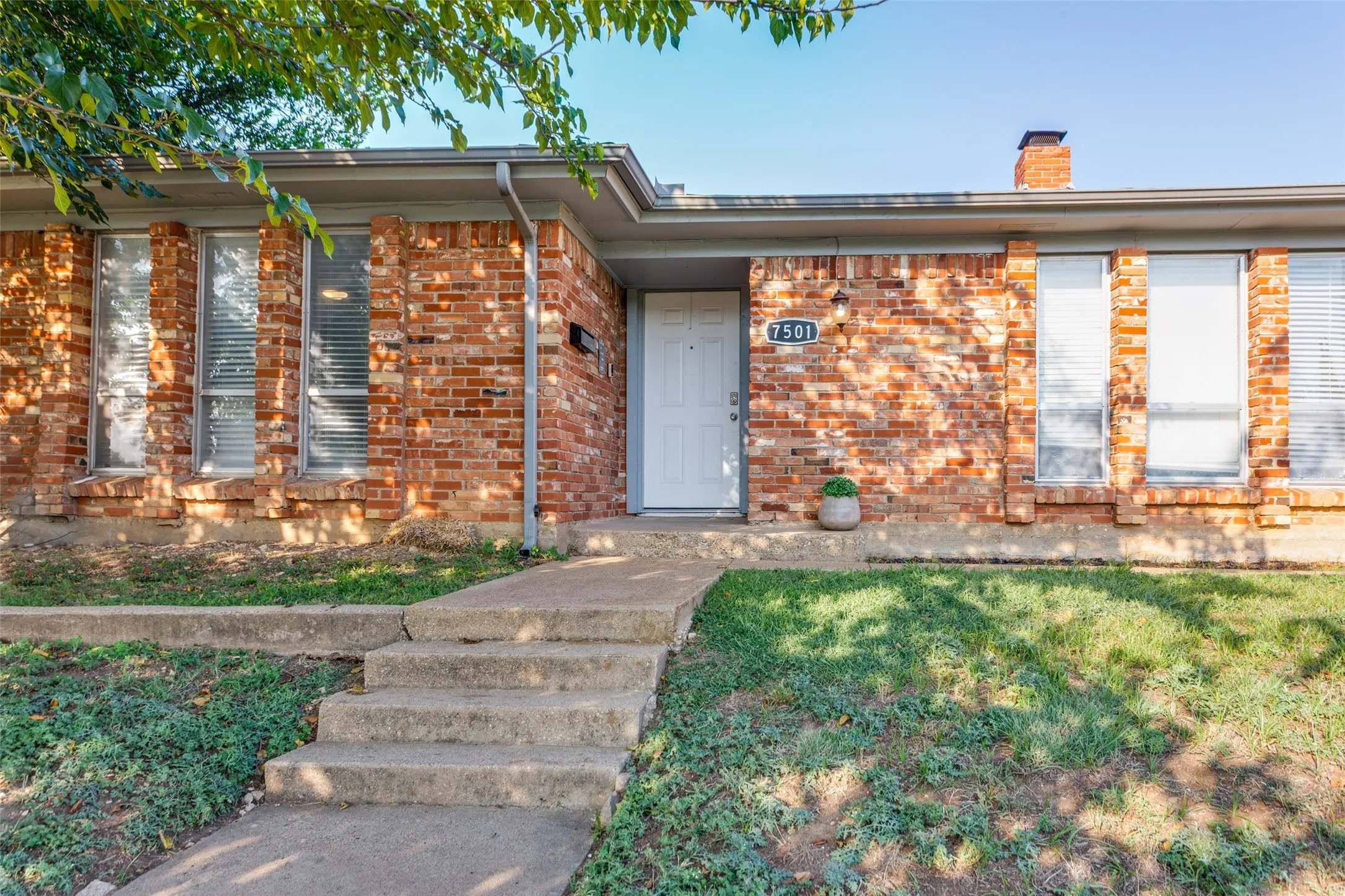 Brick exterior featuring a white entry door, multiple windows with blinds, a concrete walkway, and a partial lawn