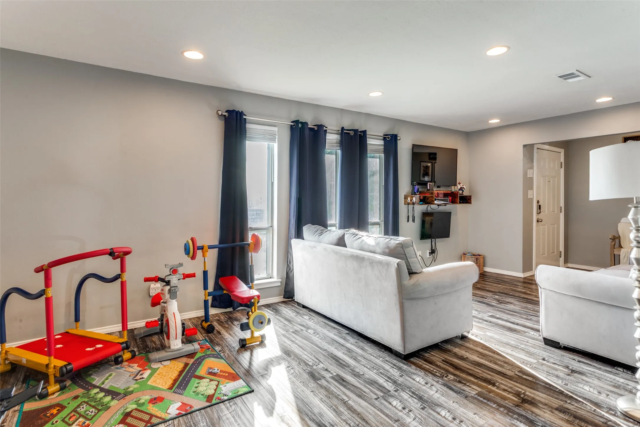 Spacious living area featuring wood-finish flooring, recessed lighting, and multiple windows with blackout curtains