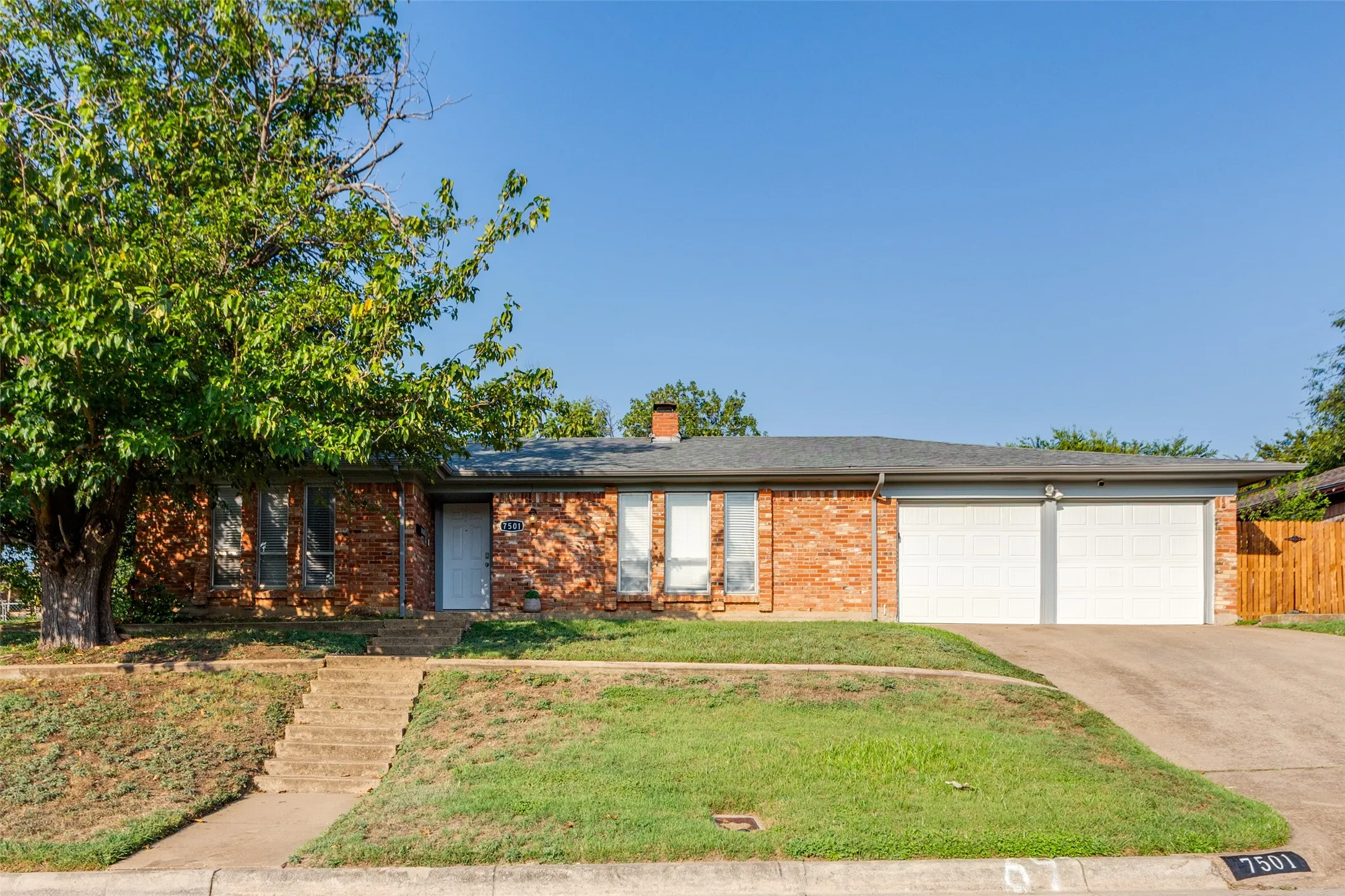 Brick-clad ranch-style residence featuring a two-car garage with white paneled doors, a mature front yard tree providing shade, concrete steps leading to the front entry, and a paved driveway
