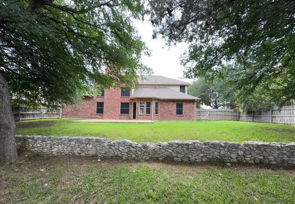 Expansive backyard featuring a stone retaining wall, mature trees, and a brick exterior home with multiple windows and a patio