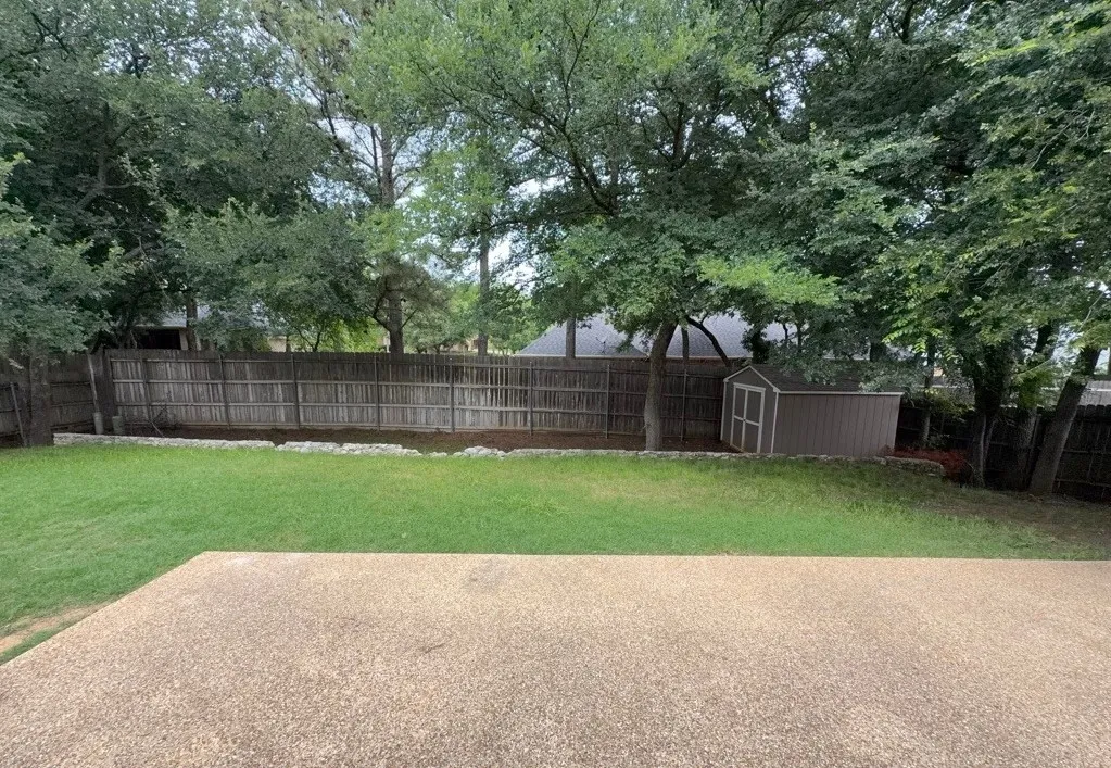 Expansive backyard featuring a concrete patio, a lush lawn, privacy fencing, mature trees, and an exterior storage shed