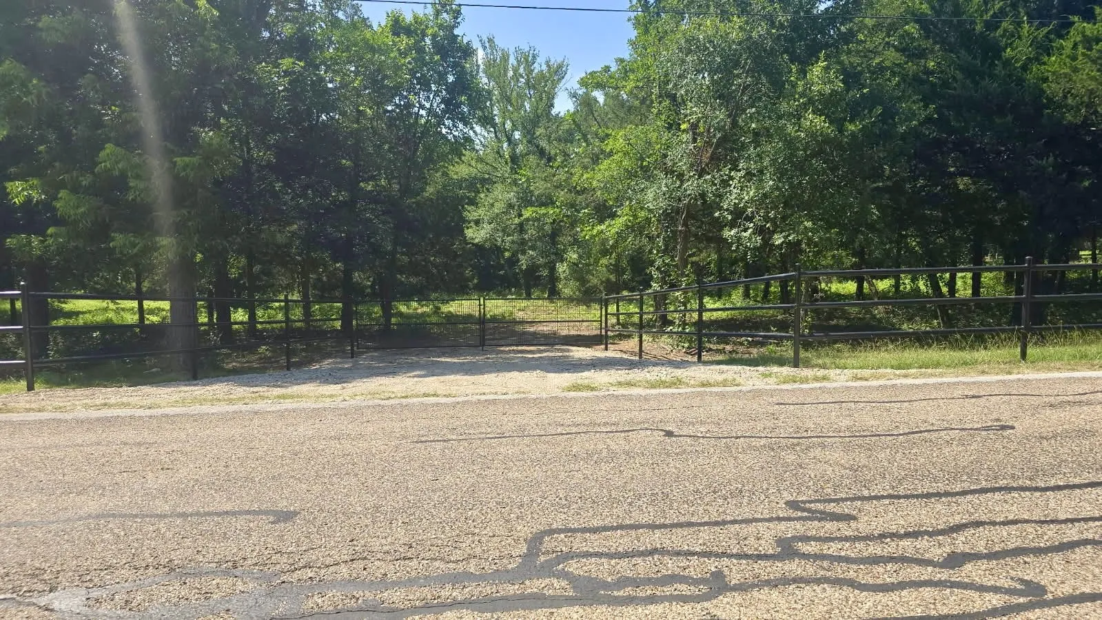 Gated property entrance with a gravel drive, bordered by mature trees and a black metal fence