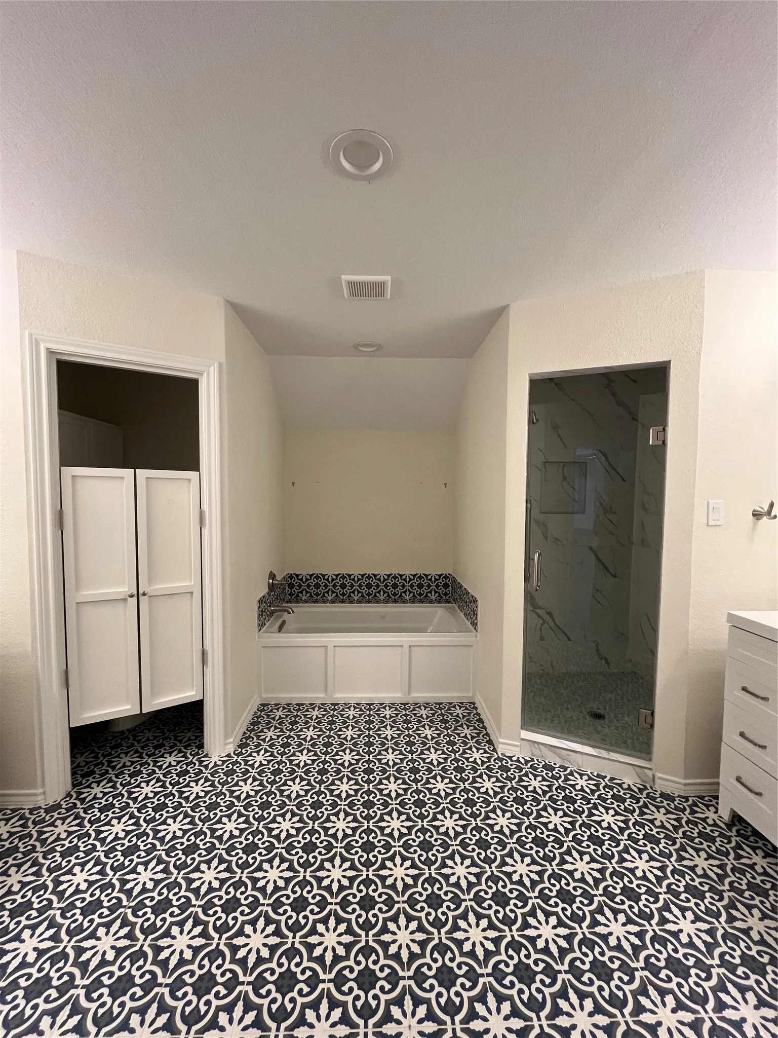 Bathroom featuring intricate patterned tile flooring, a built-in jetted tub with a tiled surround, a glass-enclosed shower with a tiled interior, a dual vanity cabinets with drawers, and bi-fold doors