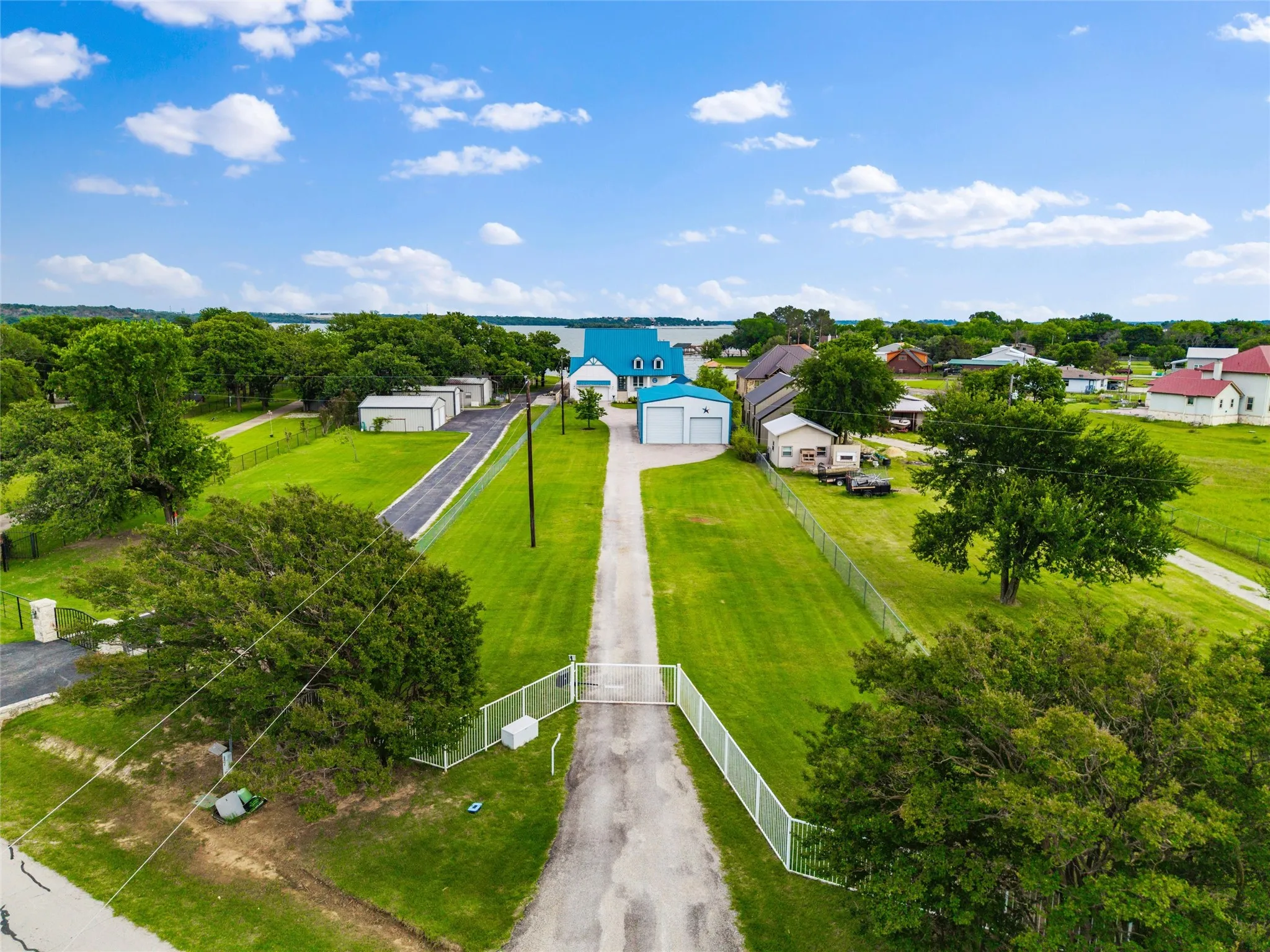 Aerial view features gated driveway access leading toward the main residence, detached garage, and guest house structures. Expansive lawn areas, perimeter fencing, mature trees, and surrounding waterfront areas are visible throughout the property.