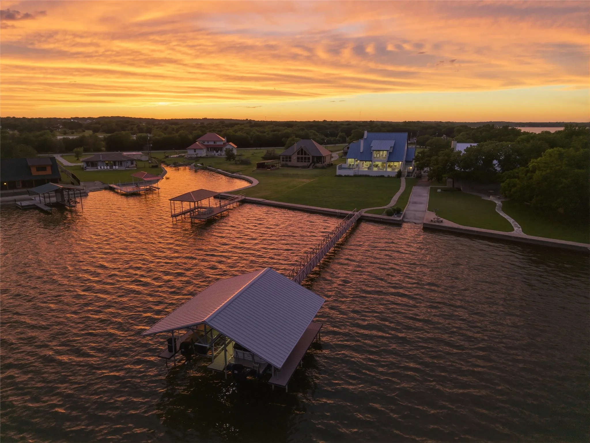 Aerial view showcases the waterfront positioning of the property with extended shoreline frontage and private dock access. Rear elevation includes expansive windows, covered outdoor living areas, and landscaped grounds extending toward the water. Concrete walkways, mature trees, and surrounding waterfront properties are visible throughout the aerial perspective.