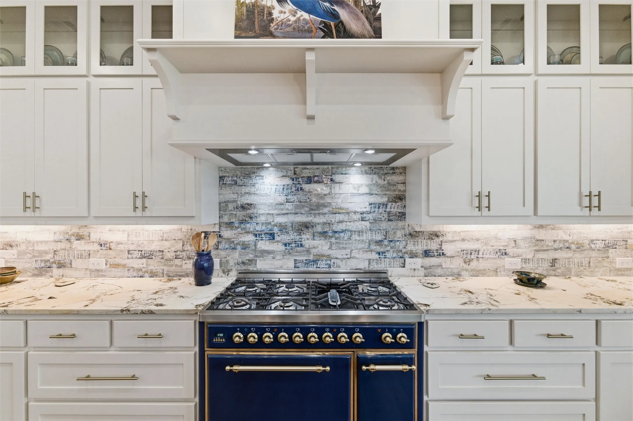 Kitchen cooking area features commercial-style gas range with double ovens, custom vent hood surround, textured tile backsplash, extensive cabinetry storage, and stone countertop surfaces extending throughout the surrounding prep areas.