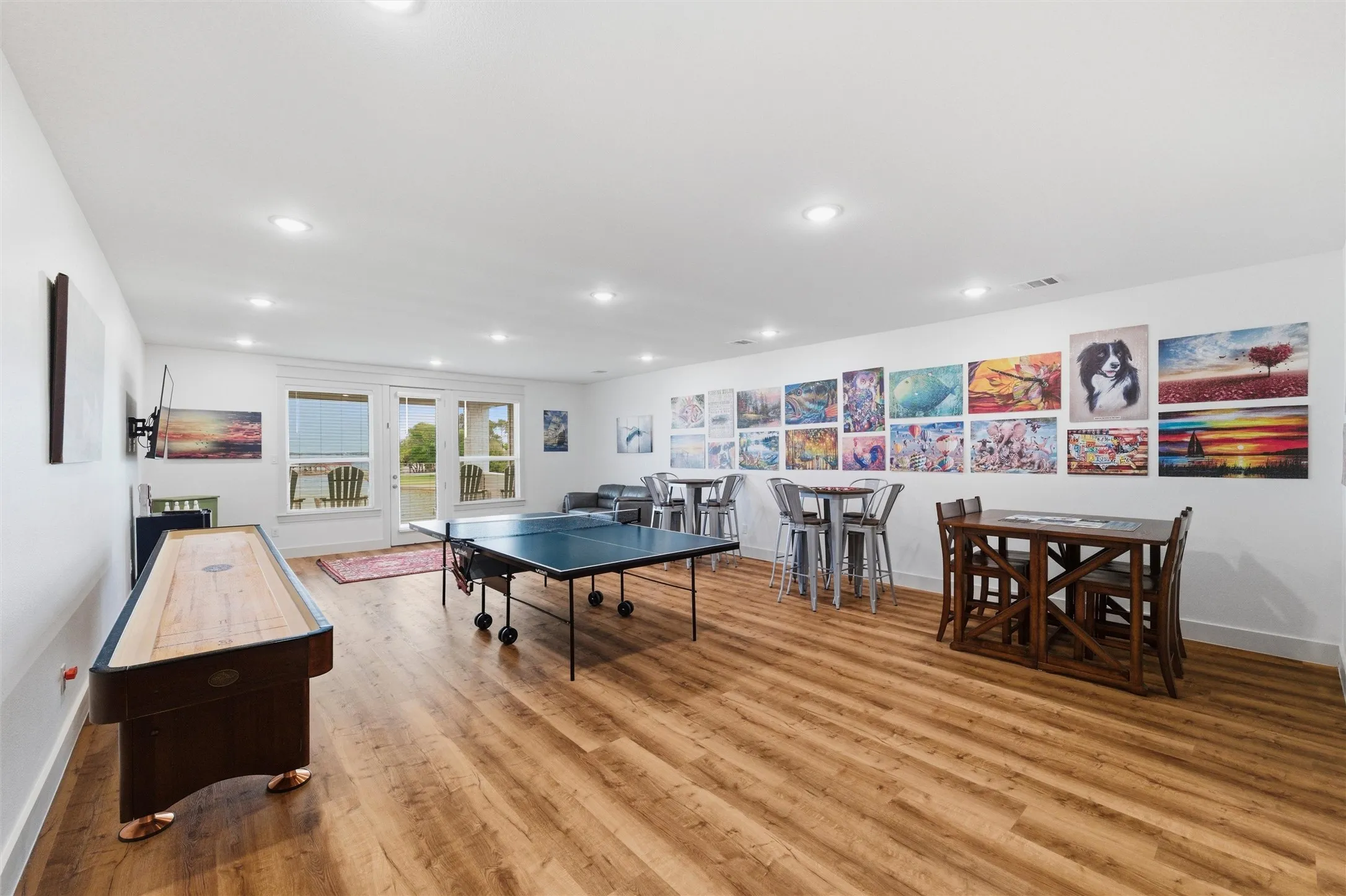 Game and entertainment room features expansive open layout with multiple gathering and recreation areas. Large windows and glass doors provide natural light and direct access to the exterior lakefront areas. Wood-look flooring and recessed lighting extend throughout the flexible entertainment space.
