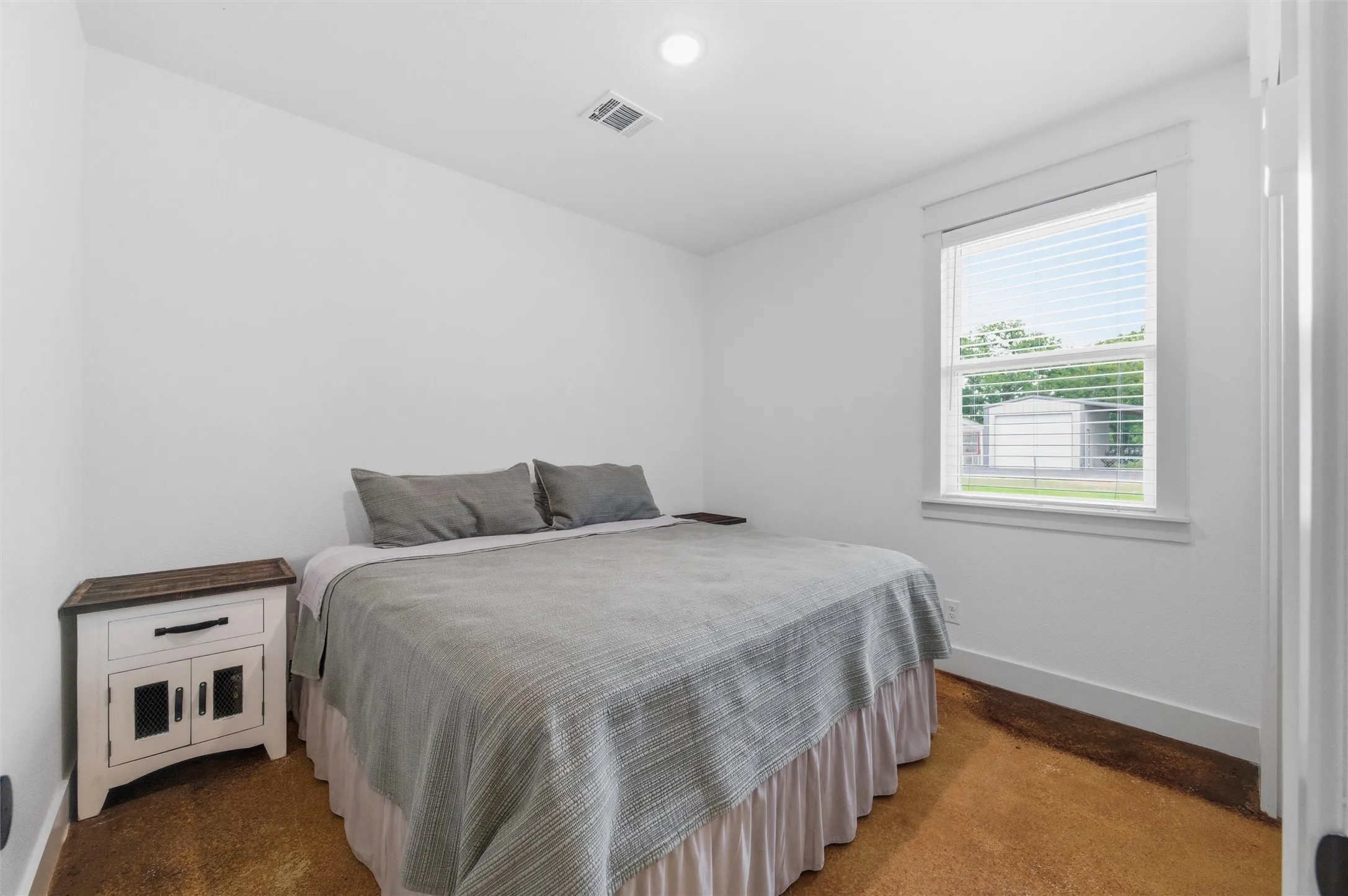 Guest bedroom features recessed lighting, large window for natural light, neutral finishes, and polished concrete flooring throughout the room.
