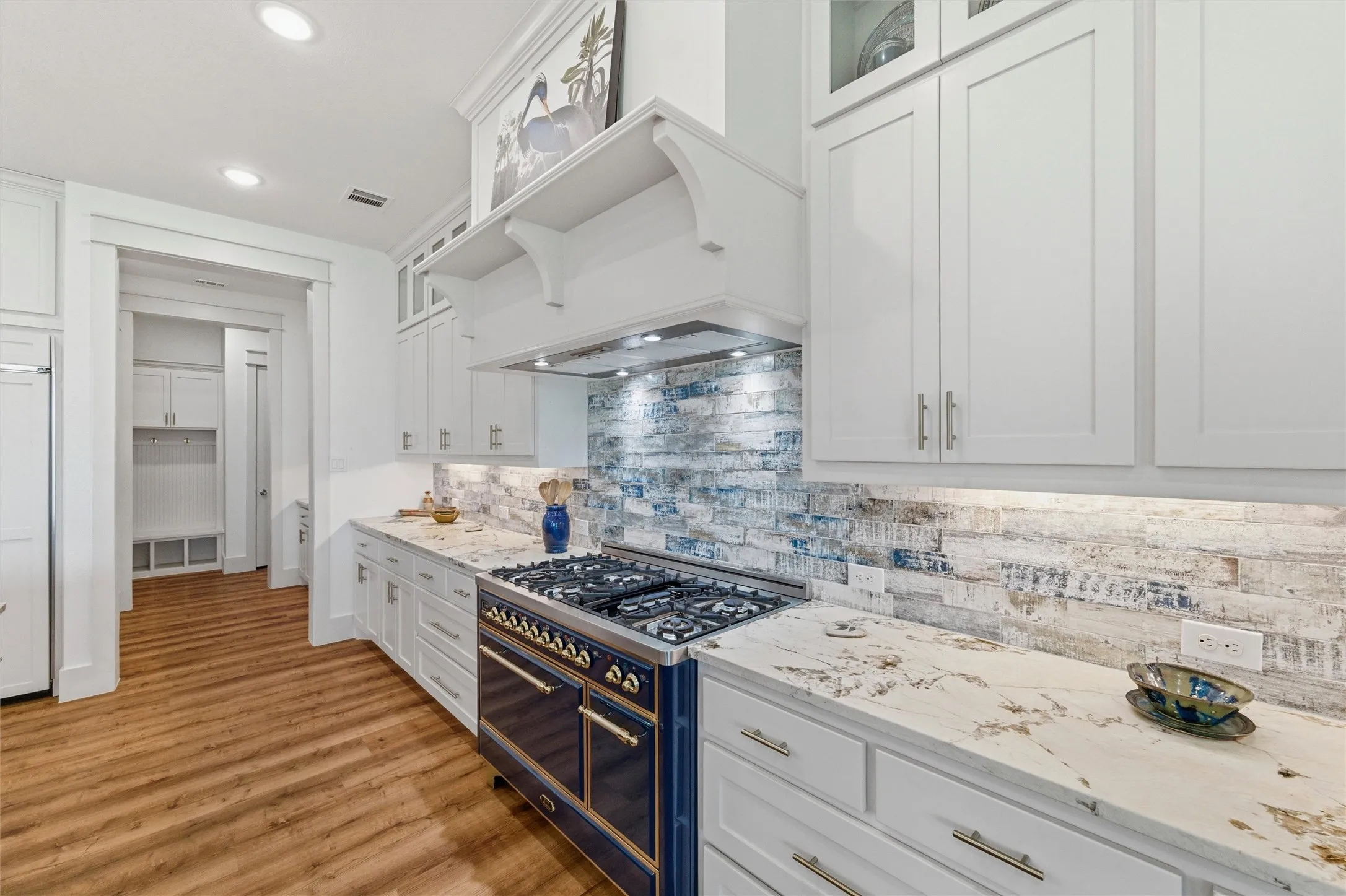 Kitchen features extensive cabinetry, glass-front upper display cabinets, stone countertops, textured tile backsplash, and a large center island with seating and undermount sink. Commercial-style gas range with double oven configuration is positioned beneath a custom vent hood enclosure. Open layout connects directly to the dining and living spaces.