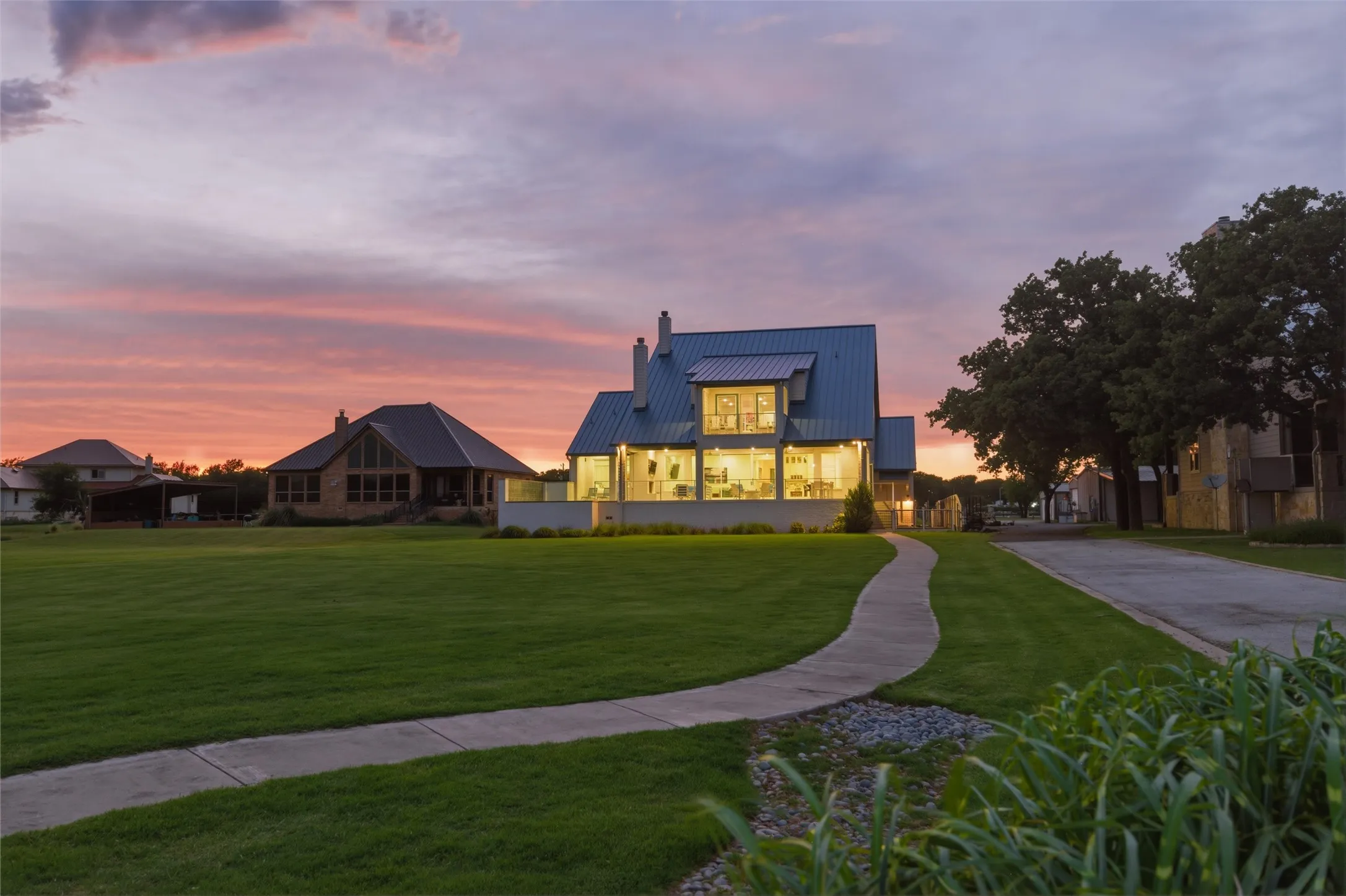 Rear exterior showcases expansive windows and glass doors overlooking the backyard and waterfront setting. Covered outdoor living areas extend along the rear elevation with landscaped lawn spaces, concrete walkways, and mature trees surrounding the property.