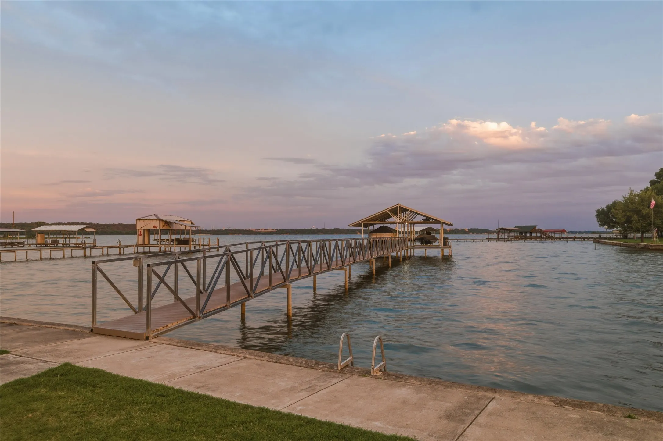 Lake view features open water views across the main body of the lake with private dock access extending from the shoreline. Covered dock structure includes walkway access with metal railing detail and covered boat slip area. Landscaped lawn areas and concrete shoreline edging border the waterfront setting.