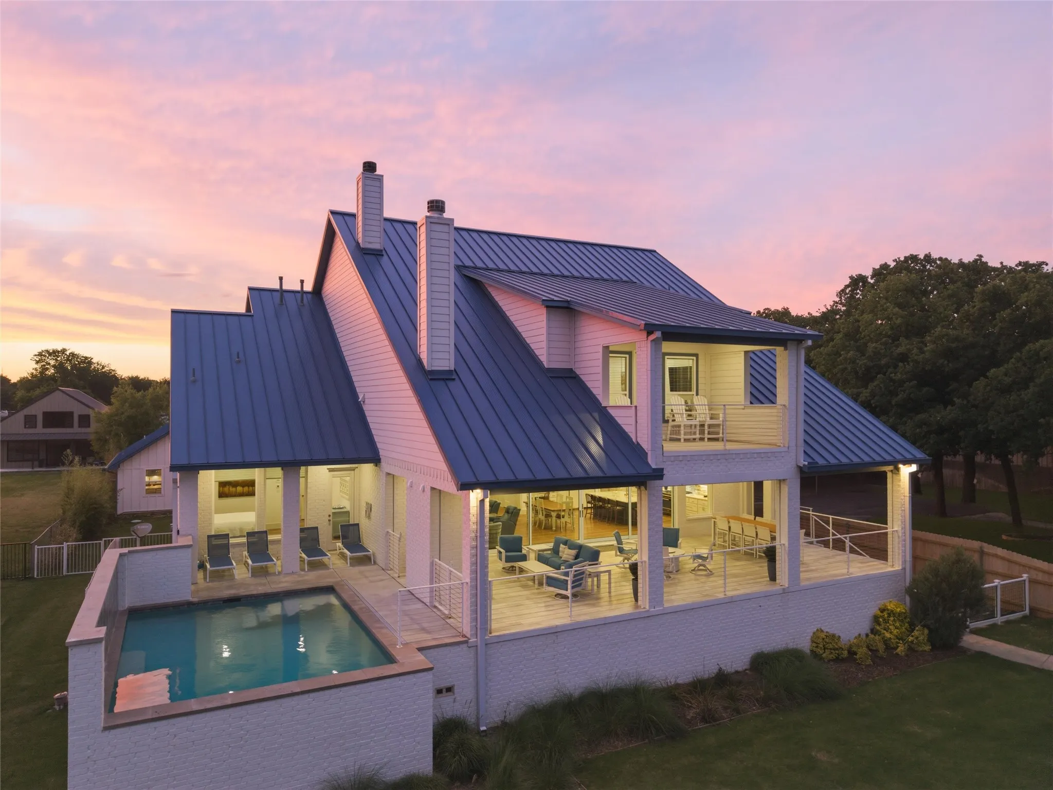 Rear exterior features multiple covered patio areas, upper balcony space with railing detail, expansive rear-facing windows, and in-ground pool overlooking the waterfront setting. Landscaped lawn areas and mature trees surround the exterior spaces.