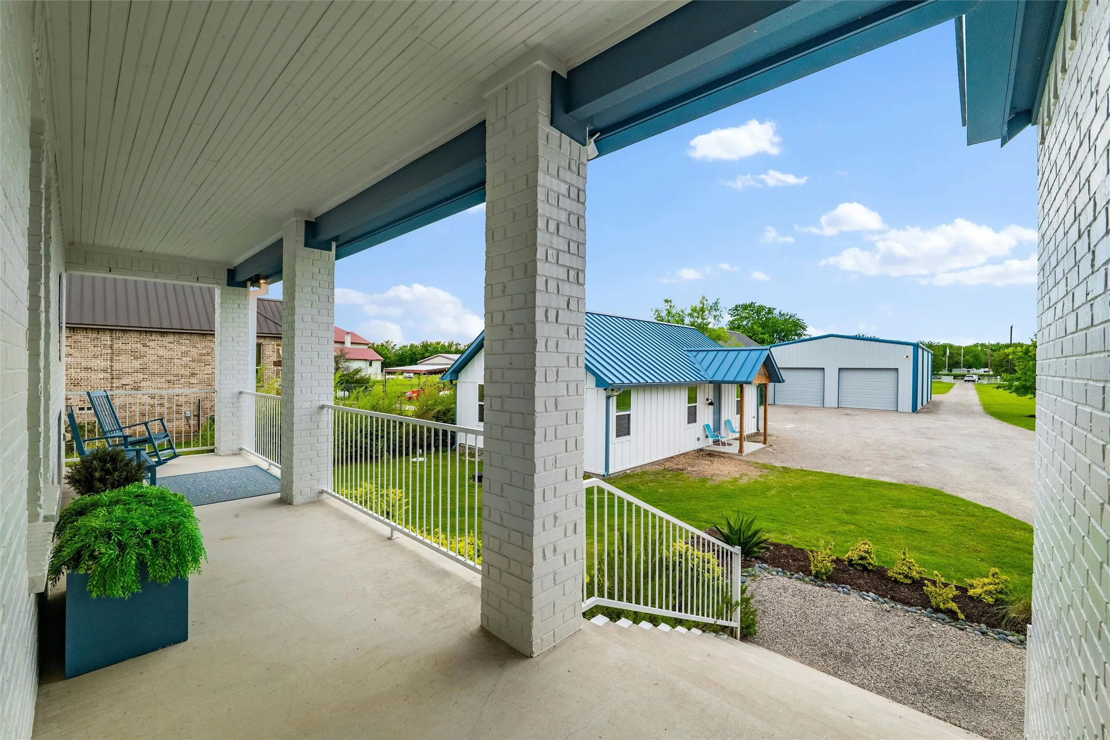 Front porch features painted brick support columns, exposed beam accents, metal railing detail, and concrete flooring throughout the covered entry area. Exterior overlooks landscaped lawn spaces, gravel driveway access, and views toward the detached guest house and garage structures.