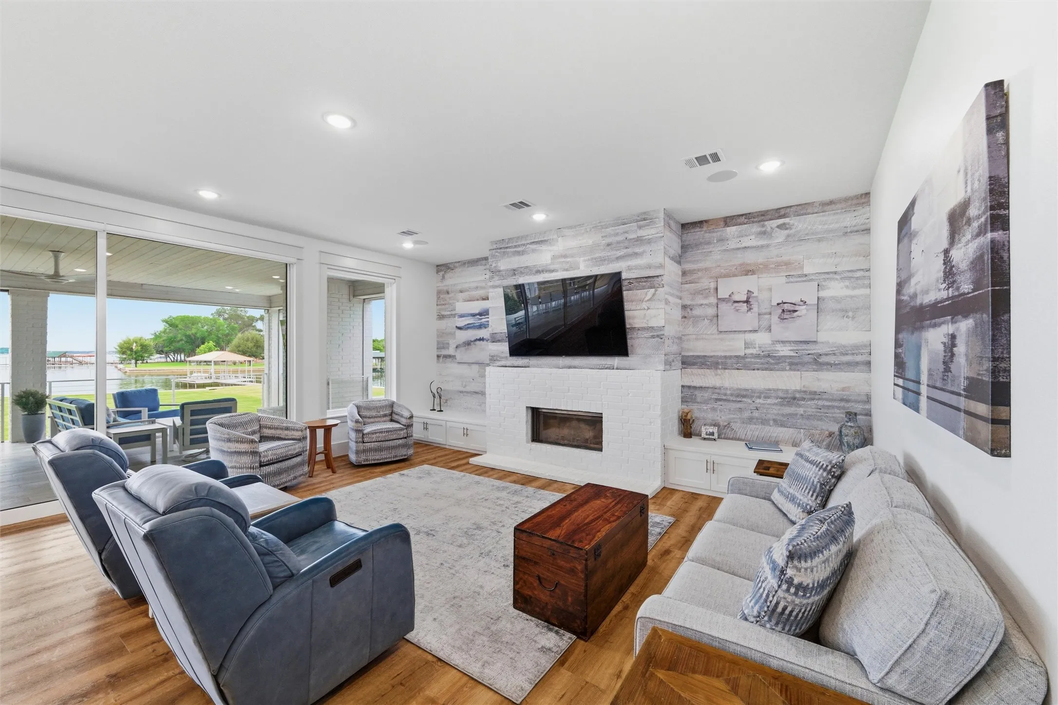 Living room features recessed lighting, built-in cabinetry, painted brick fireplace surround with raised hearth, and wood accent wall with mounted television area. Expansive sliding glass doors and large windows provide natural light and direct views toward the covered patio and waterfront setting. Open-concept layout connects seamlessly to the dining and kitchen areas with wood-look flooring extending throughout.