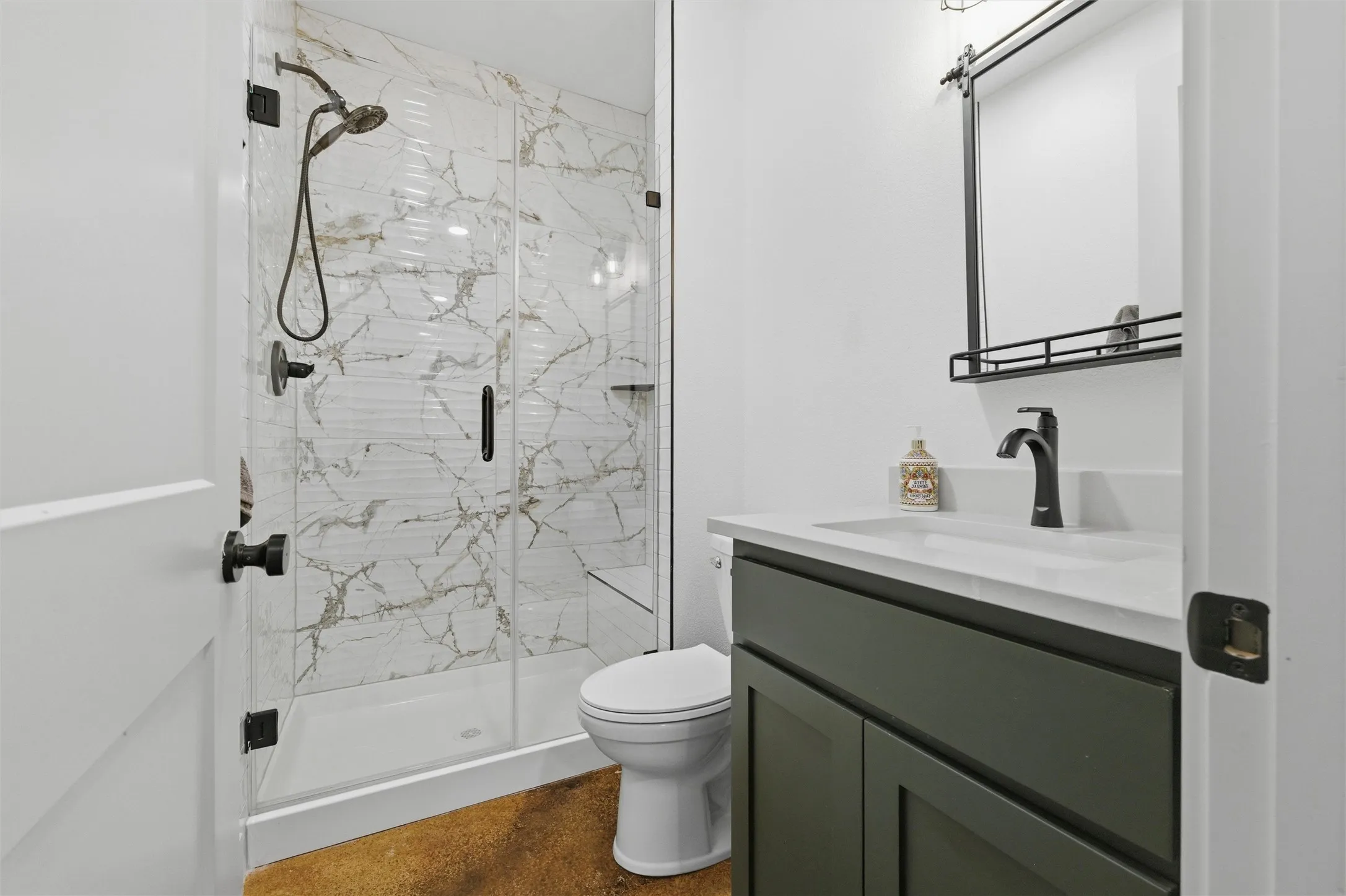 Guest bathroom features walk-in glass shower with full-height tile surround, modern black fixtures, sleek vanity with quartz countertop, and polished concrete flooring throughout.
