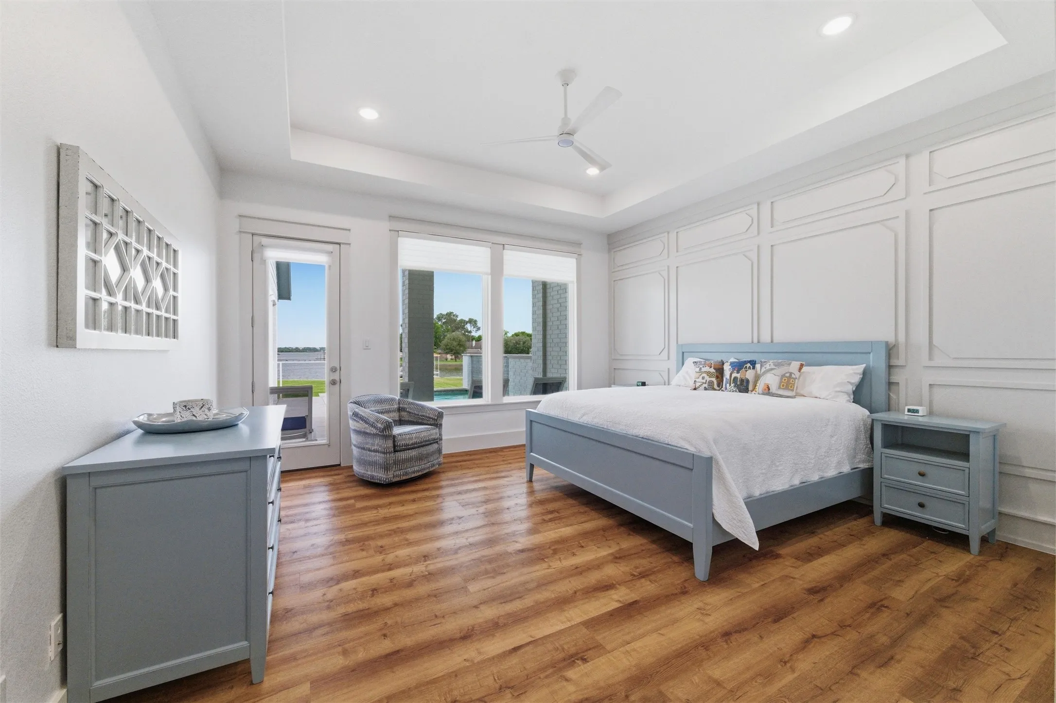 Primary bedroom features tray ceilings with decorative wall trim detailing, modern ceiling fan, recessed lighting, and large windows with glass door access to the exterior. Spacious layout overlooks the waterfront setting with wood-look flooring extending throughout the room.