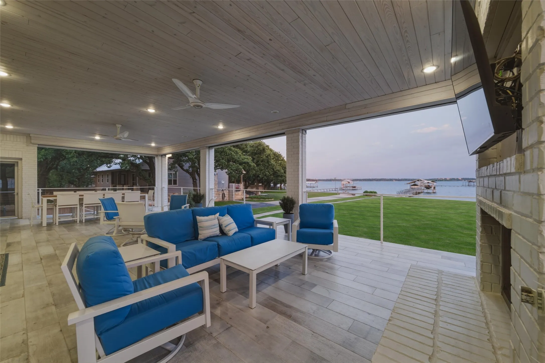 Covered patio overlooks the waterfront setting with expansive open-air views across the lawn, dock access, and surrounding shoreline. Exterior fireplace with mounted television niche creates an additional outdoor gathering area beneath the covered patio structure.
