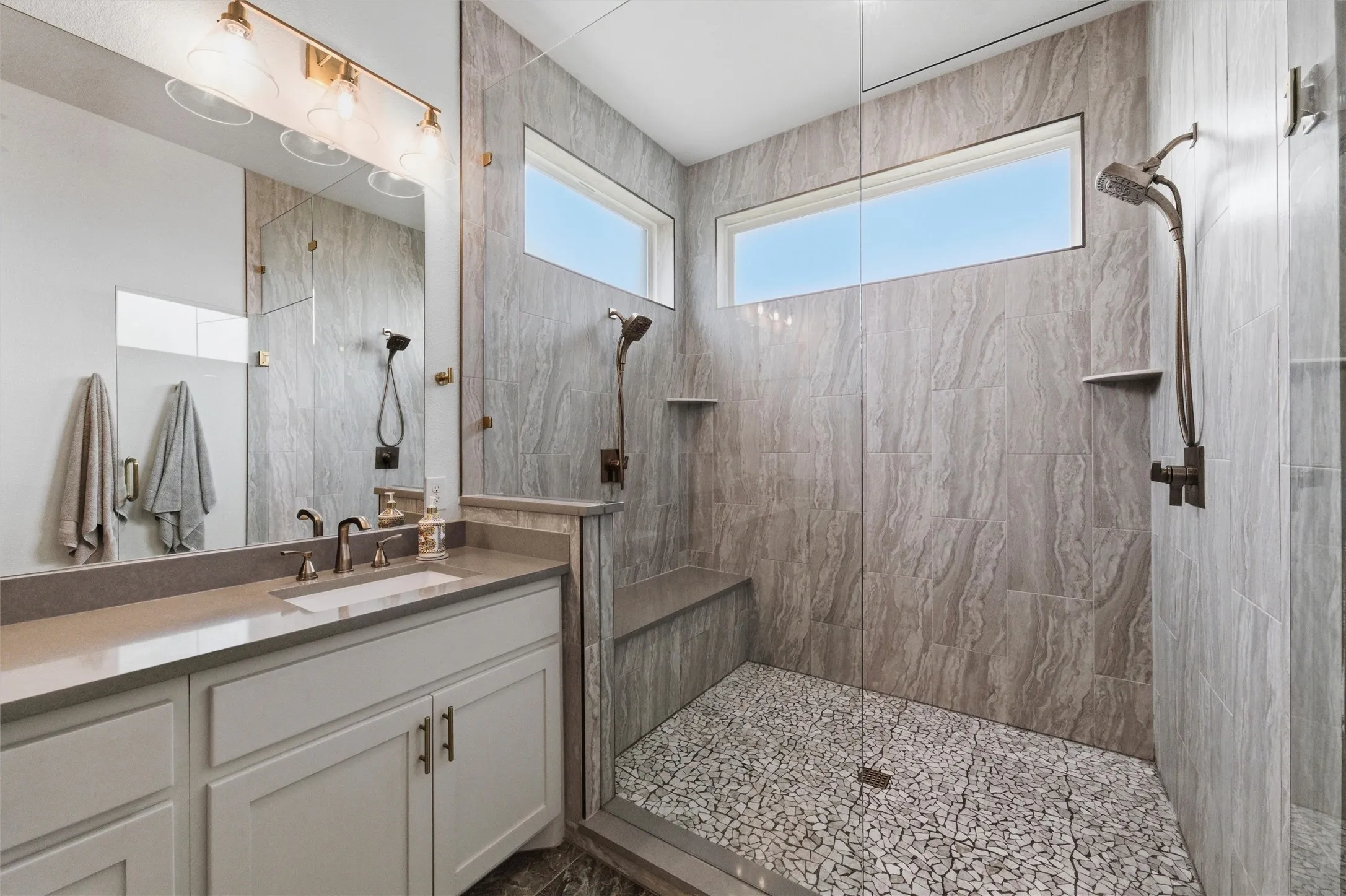 Primary bathroom showcases oversized walk-in shower with full-height tile surround, pebble-style shower flooring, built-in bench seating, and dual vanity layout with extensive storage and modern fixtures throughout.
