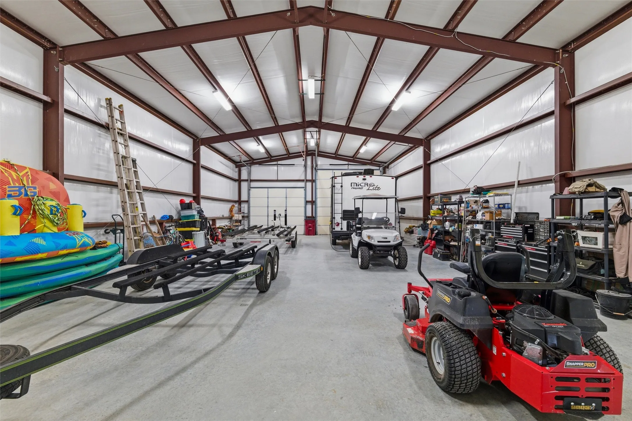 Garage interior features exposed steel beam construction, insulated wall and ceiling panels, overhead lighting, concrete flooring, and large open layout suitable for vehicle storage, equipment, trailers, and recreational items.