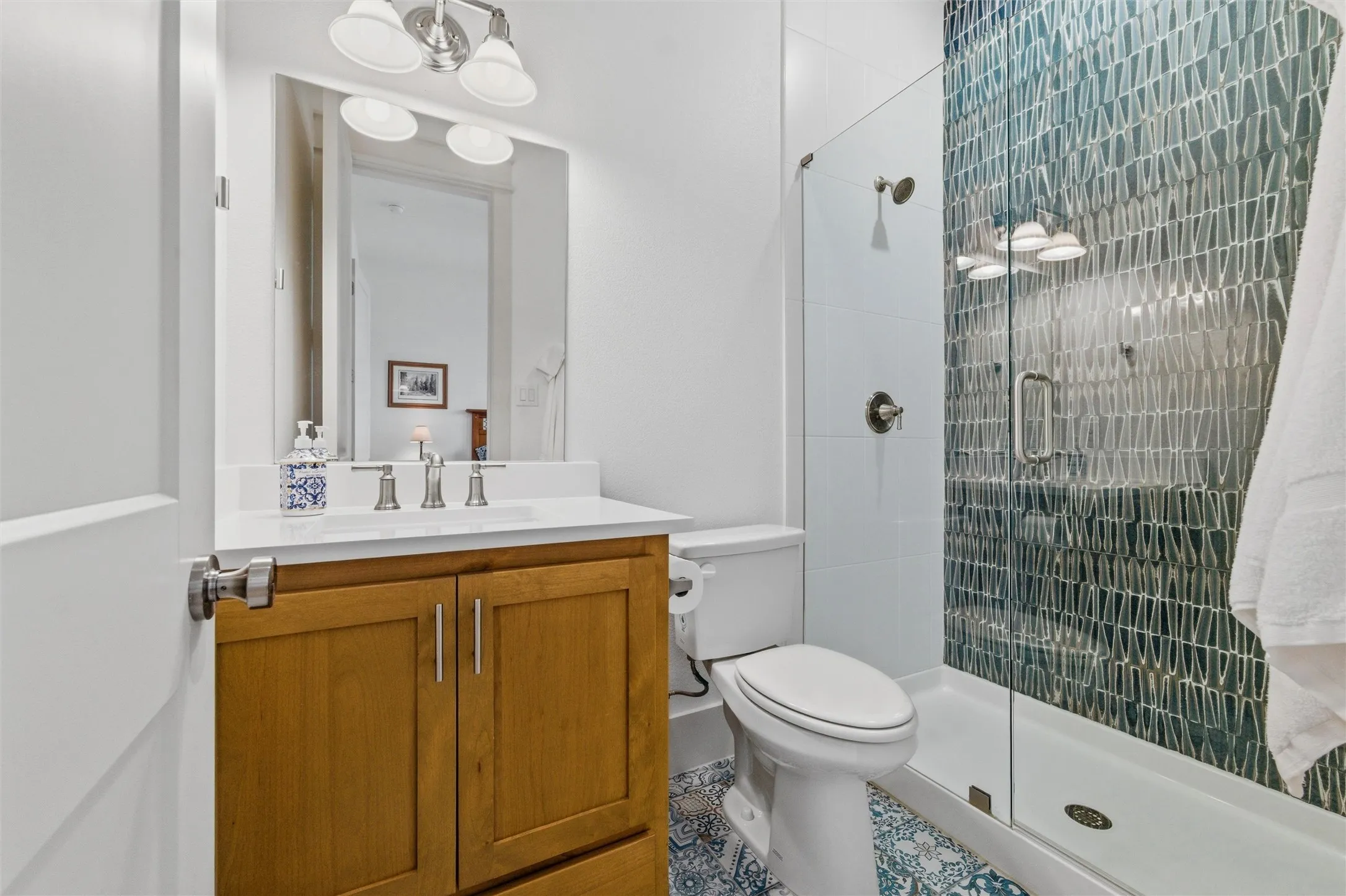 Secondary bathroom features glass-enclosed walk-in shower with decorative full-height tile accent wall, wood-tone vanity cabinetry, quartz-style countertop, framed mirror, and patterned tile flooring throughout.