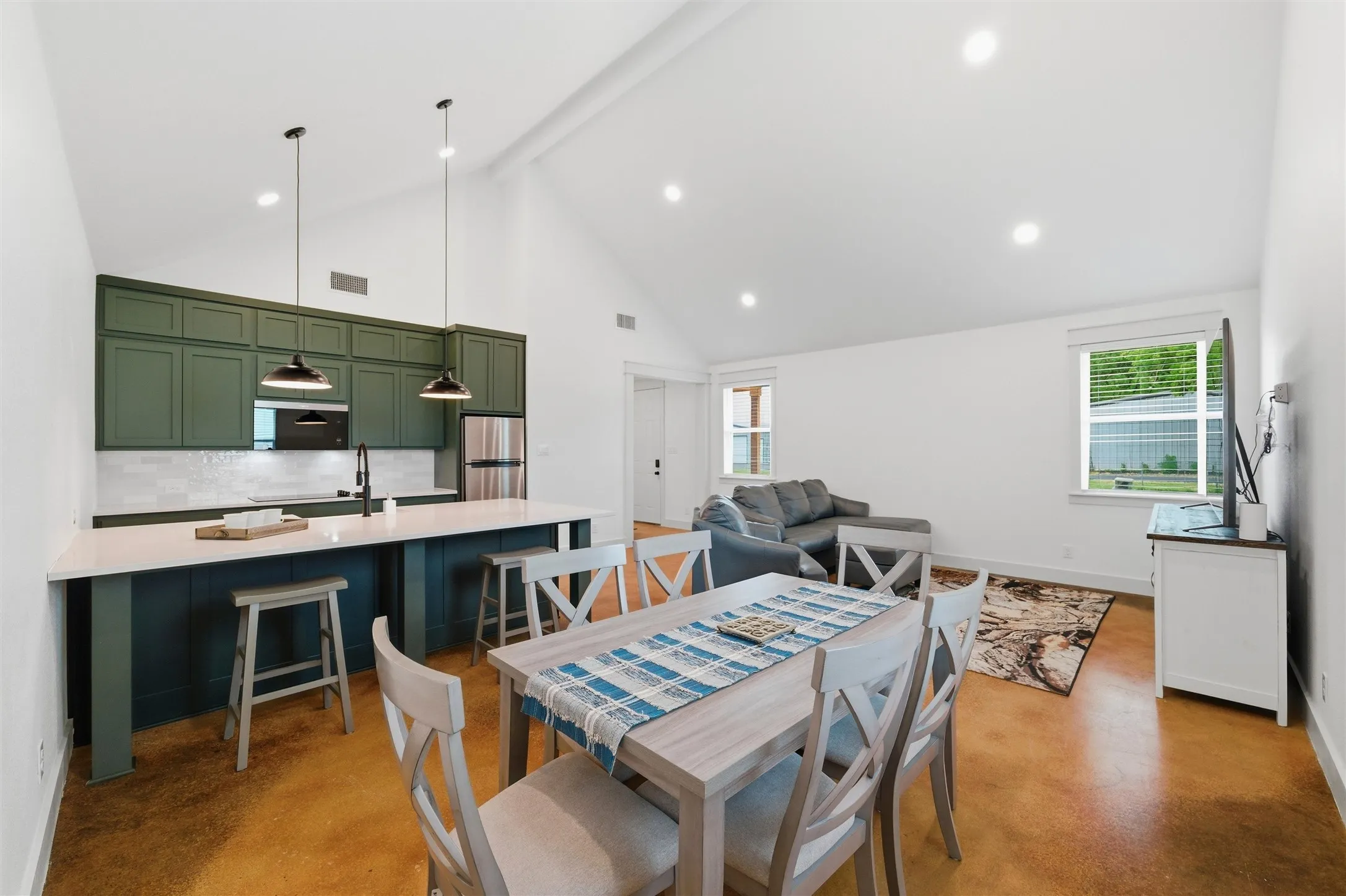 Guest house open-concept layout connects the kitchen, dining, and living areas with oversized island seating, large windows, recessed lighting, and polished concrete flooring extending throughout the interior space.