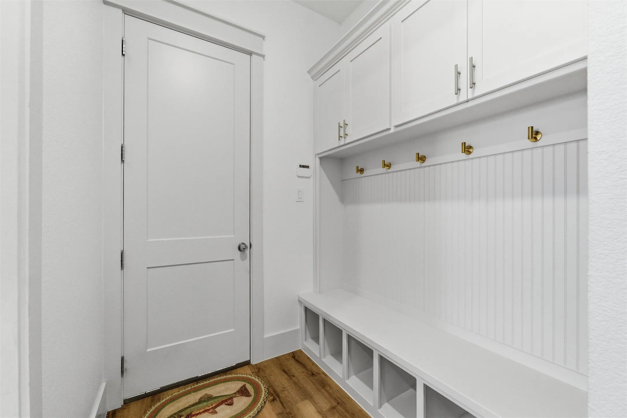 Mudroom features built-in bench seating, cubby storage, wall-mounted hooks, upper cabinetry, and wood-look flooring throughout the space. Functional storage layout provides organization and everyday utility near the interior entry area.