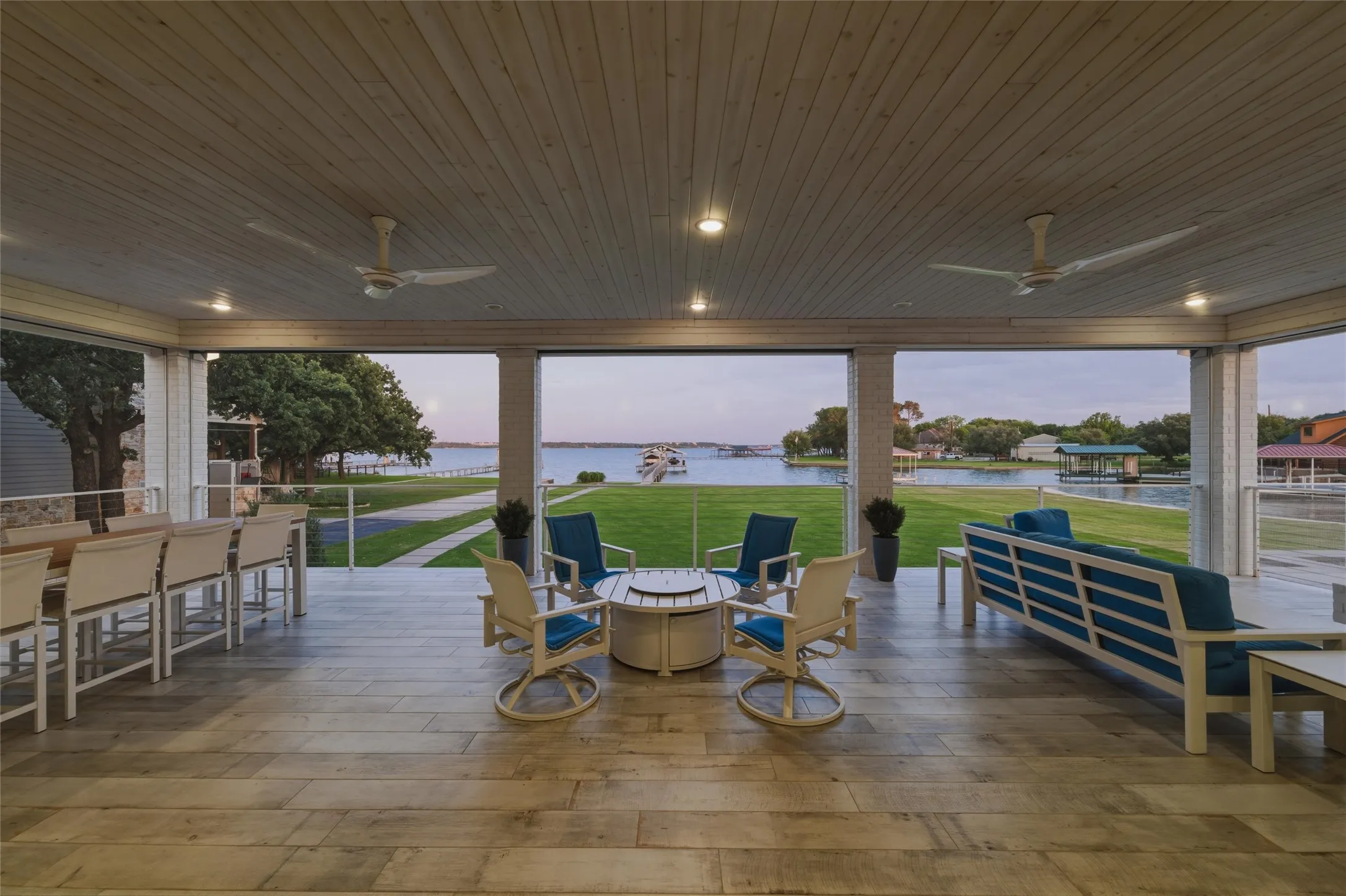 Covered patio features painted brick support columns, recessed lighting, mounted ceiling fans, cable railing detail, and large-format tile flooring throughout the outdoor living area. Open layout overlooks the lawn spaces, private dock access, and surrounding waterfront views.