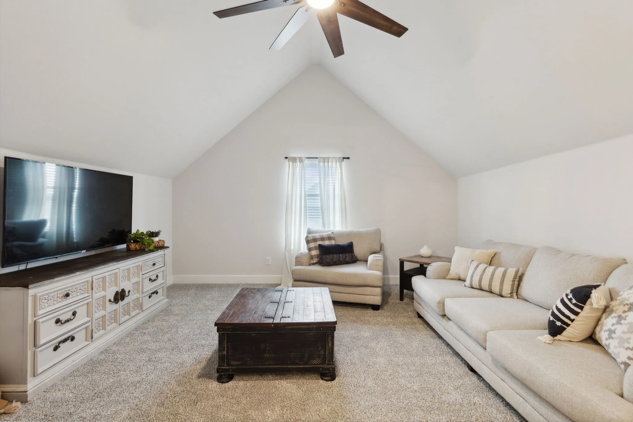 Spacious room featuring vaulted ceilings, light-toned carpeting, a window with horizontal blinds and white drapes, and a ceiling fan with integrated lighting