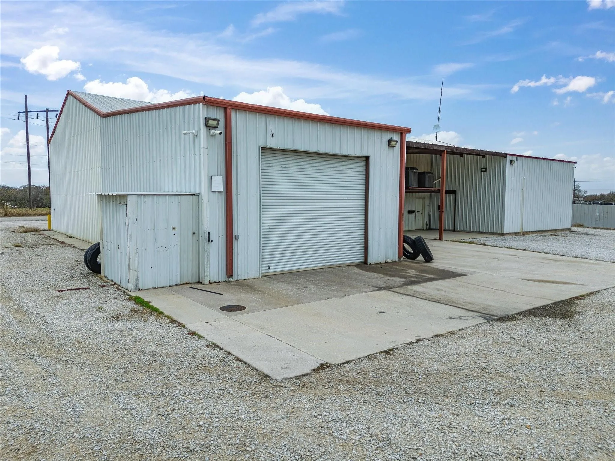 Industrial metal building featuring a roll-up door, exterior lighting fixtures, and a gravel lot with concrete pads