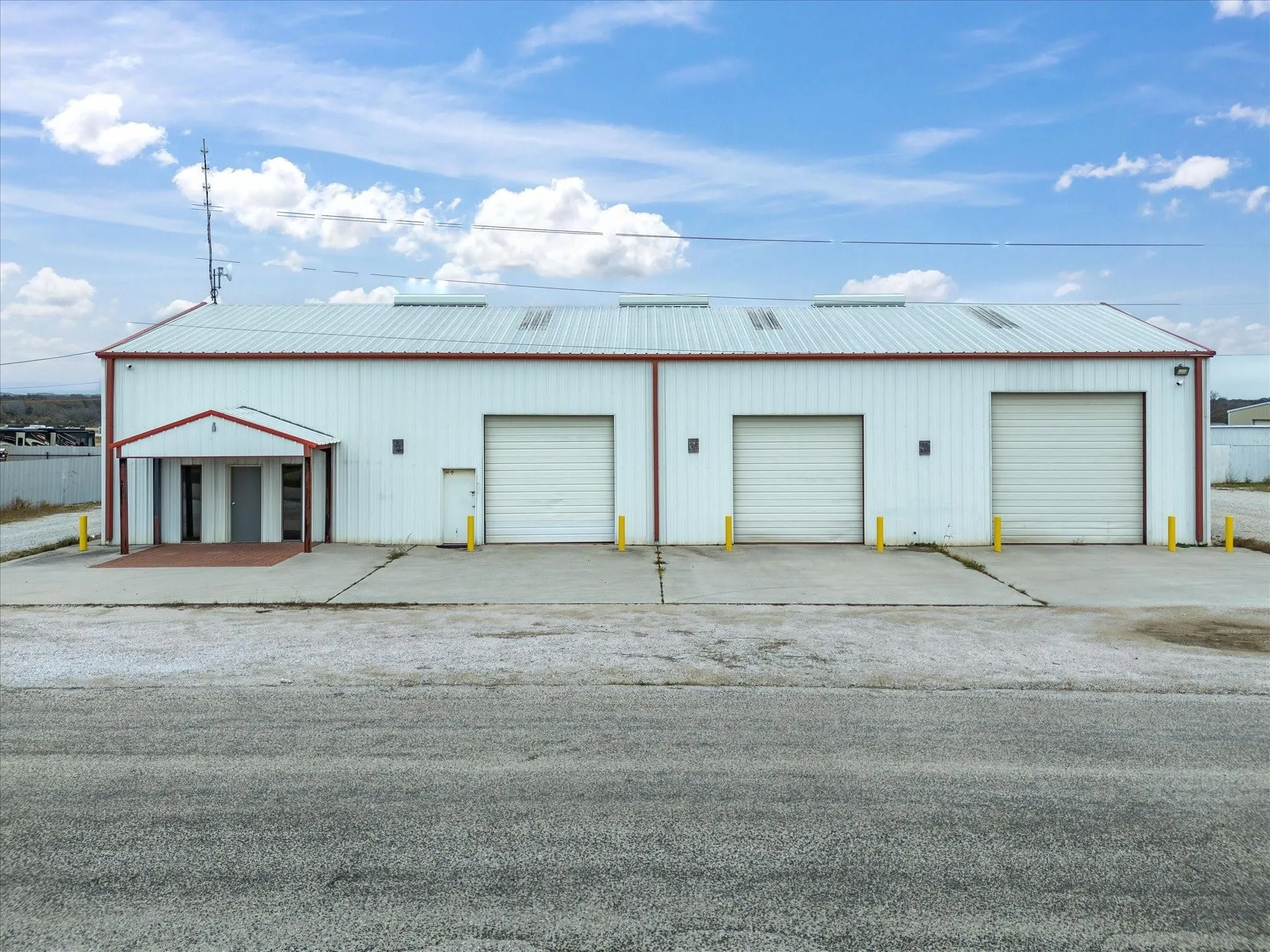 Commercial building featuring a white metal facade with red trim, three roll-up doors, a covered entry porch, and concrete paving