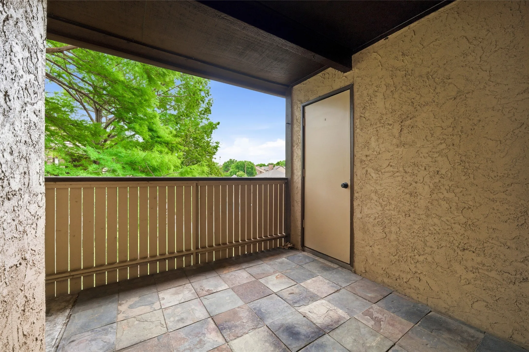 Private balcony featuring multi-toned square tile flooring, wood-slat railing, a textured stucco exterior wall, and a solid-panel door with dark hardware