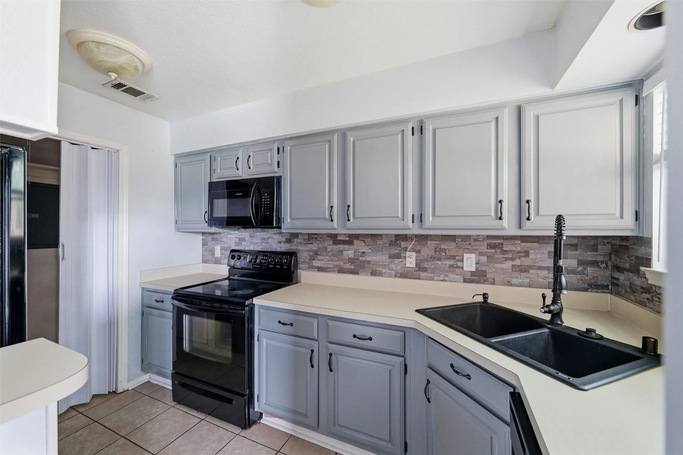 Kitchen featuring light gray cabinetry, a stone tile backsplash, light-toned countertops, tile flooring, and a black double basin sink with a matching faucet