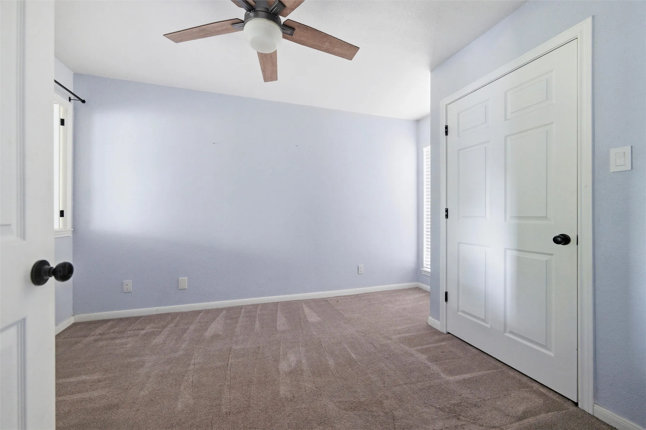 Carpeted interior space featuring light blue wall paint, a ceiling fan with integrated lighting, white trim, and a white six-panel door with a black knob