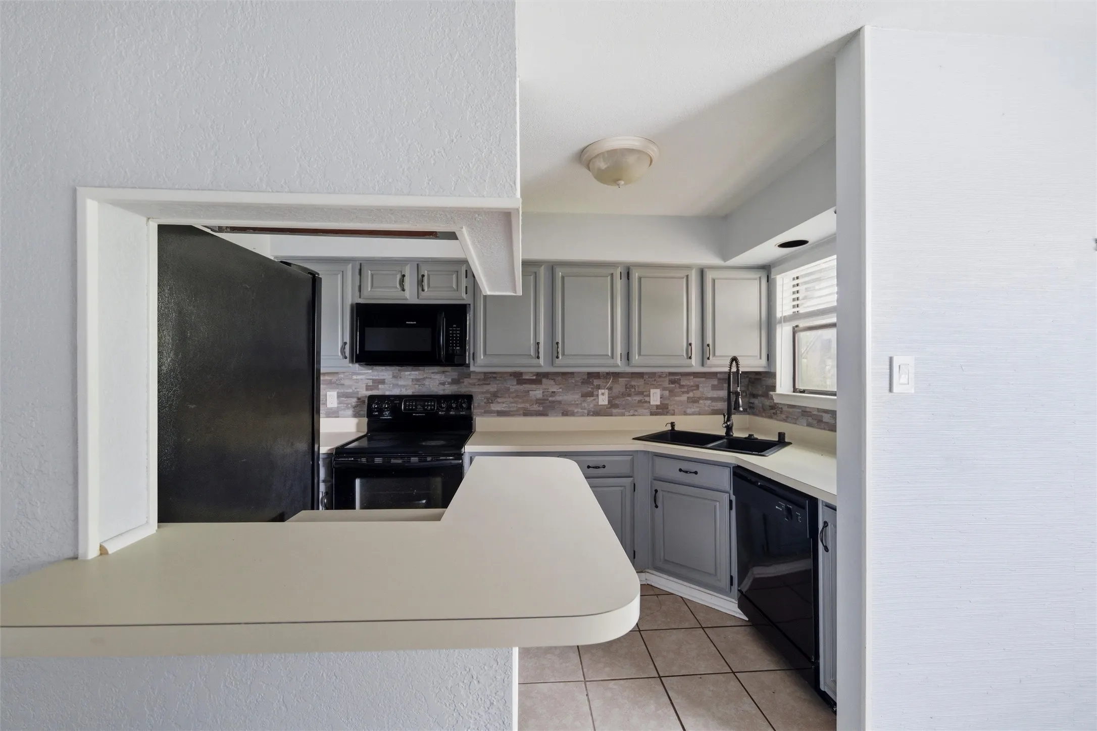 Kitchen featuring a pass-through window, light gray cabinetry, black appliances, a tile backsplash, and light-toned countertops