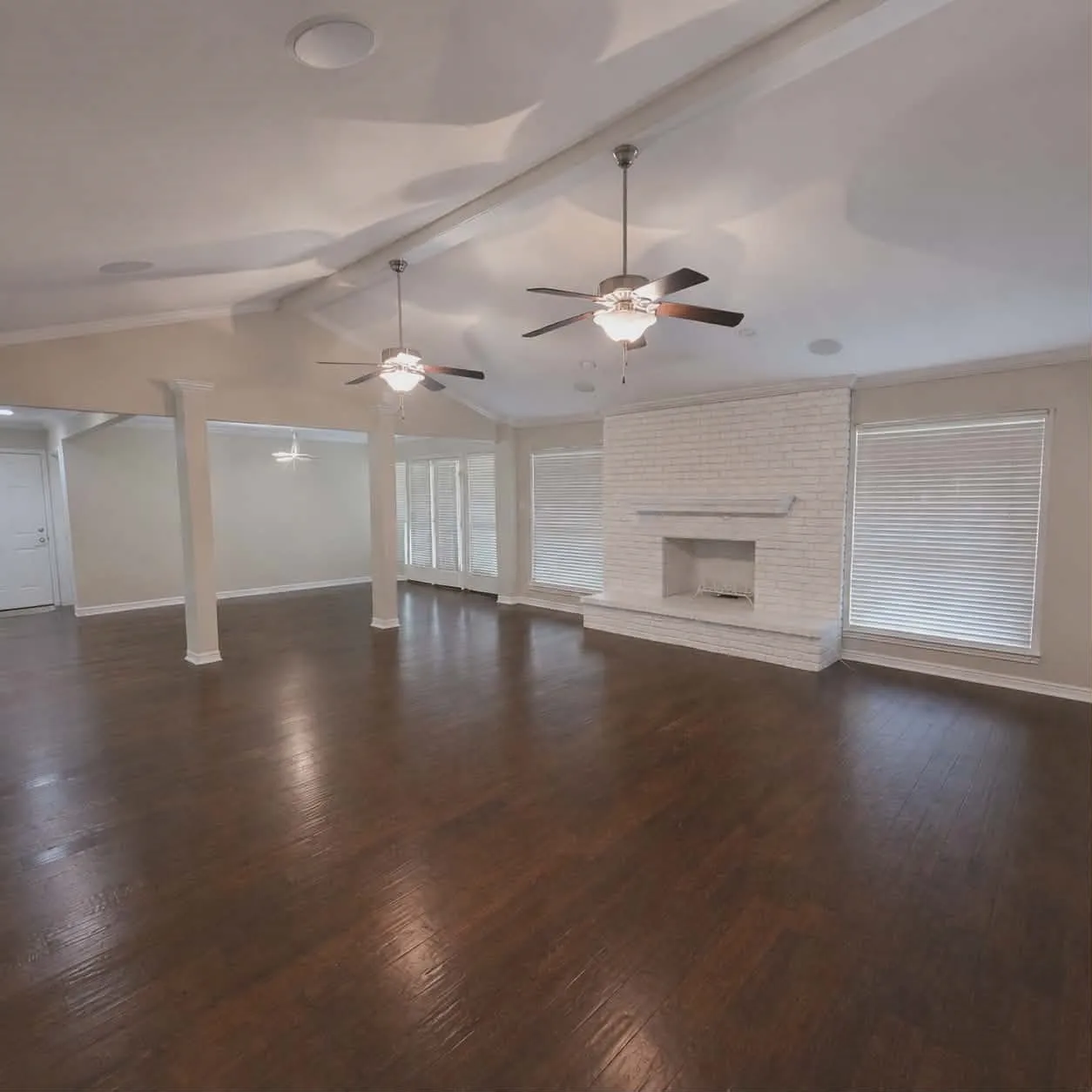 Spacious living area featuring rich wood-finish flooring, a white-painted brick fireplace with mantel, and multiple windows with blinds-This photo was taken before family moved in