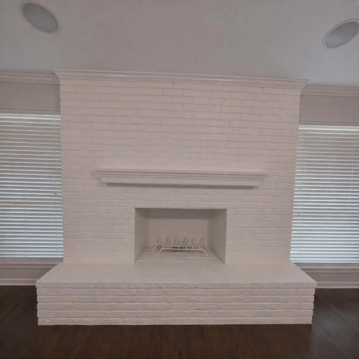 Expansive white-painted brick fireplace featuring a hearth and mantel, complemented by dark wood-finish flooring, crown molding, and recessed ceiling speakers- This photo was taken before family moved in