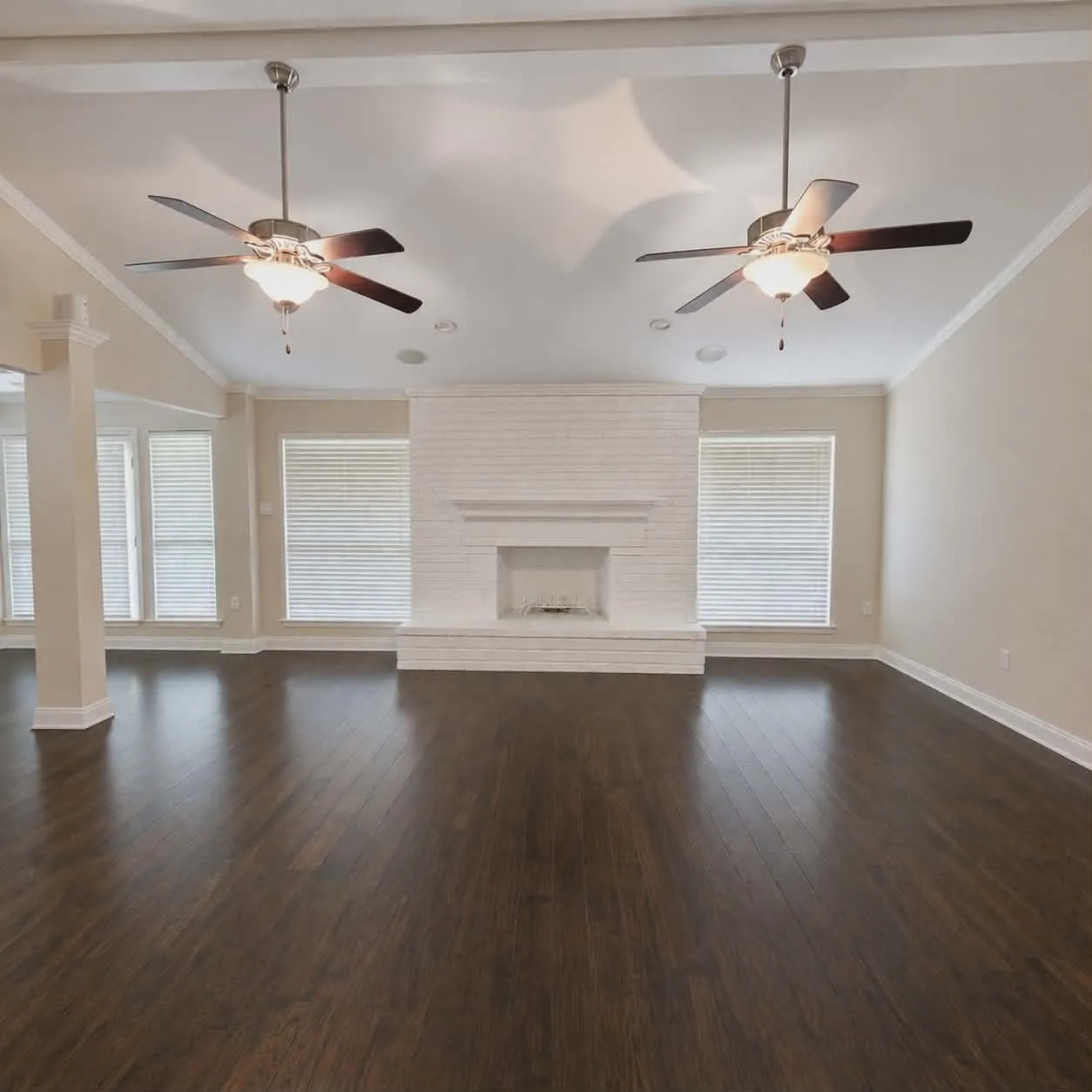 Spacious living area featuring dark wood-finish flooring, a white brick fireplace, multiple windows with blinds, and two ceiling fans with integrated lighting-This photo was taken before family moved in