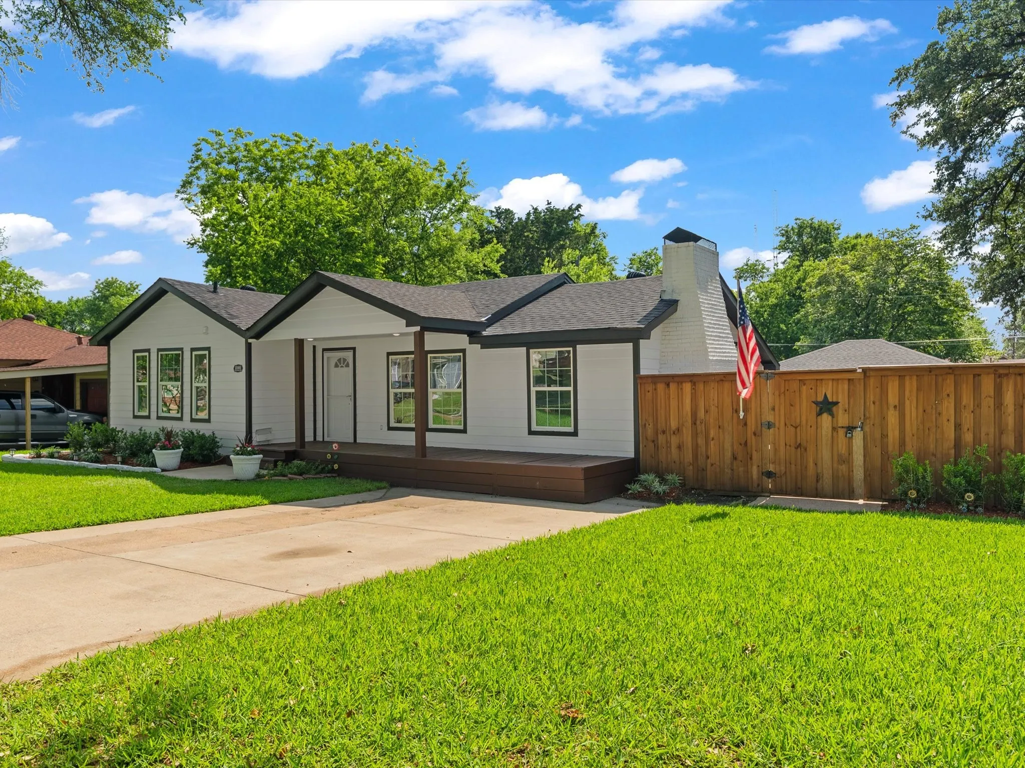 White siding exterior with a dark roof and trim, featuring a covered porch with wood-finish flooring, a concrete driveway, and a wood privacy fence