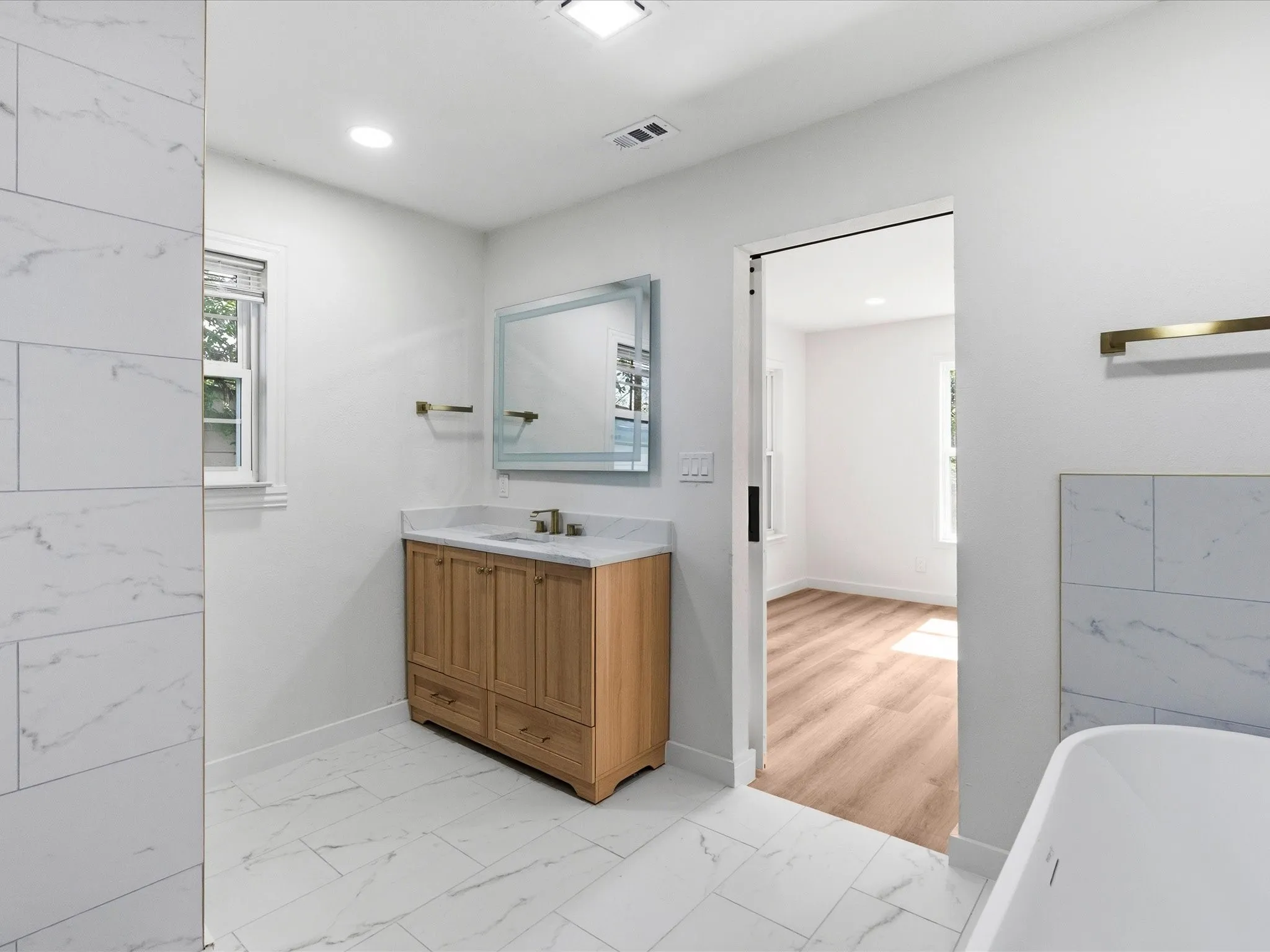 Bathroom featuring marble-look tile flooring, a wood-finish vanity with a stone countertop, a rectangular mirror, and a white soaking tub