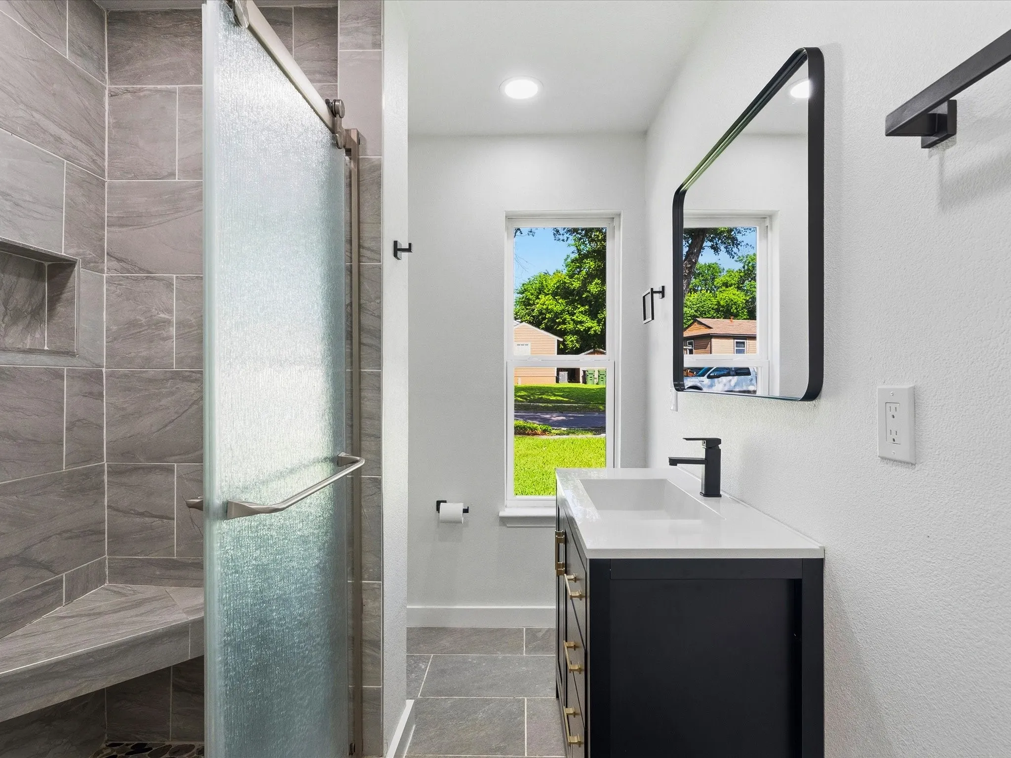 Bathroom featuring a glass-enclosed shower with a built-in bench, gray tile flooring and shower surround, a vanity with a white countertop and black cabinetry, and a black-framed mirror
