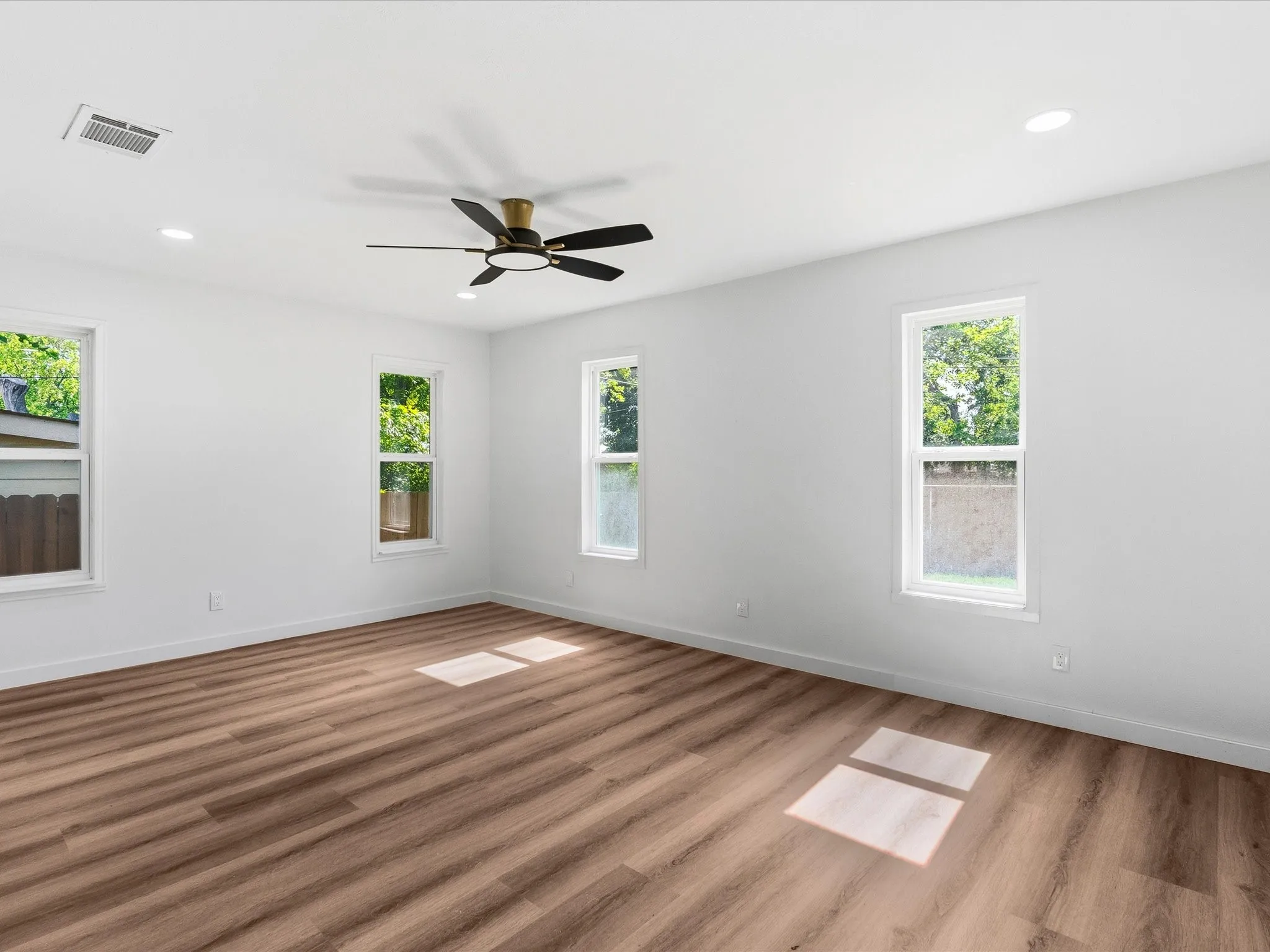 Spacious room featuring wood-finish flooring, multiple windows, a ceiling fan with integrated lighting, recessed lighting, and white walls