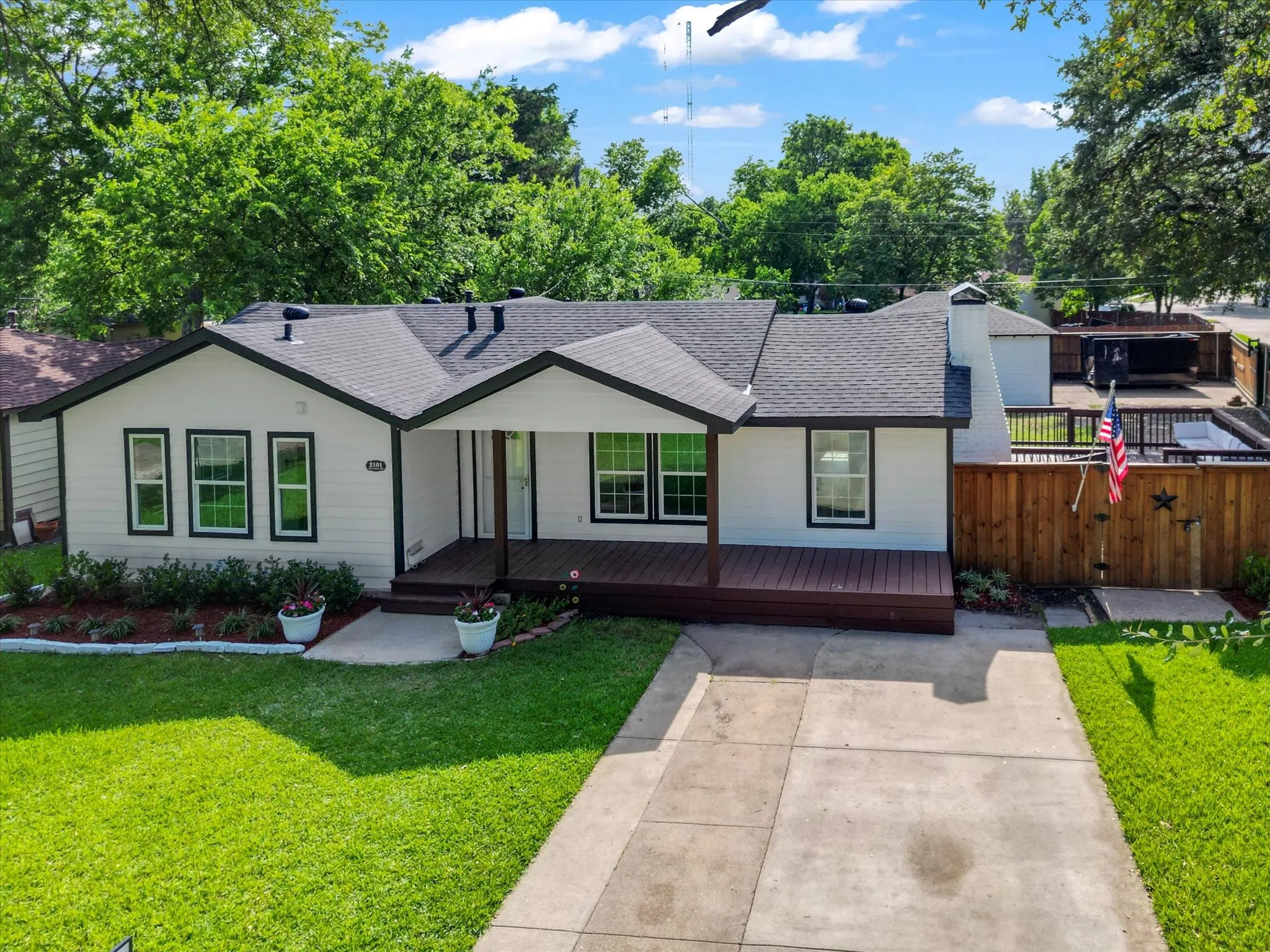Contemporary ranch-style home featuring white siding with black trim, a dark shingle roof, an elevated wood-finish front porch, and a fenced side yard with a patio