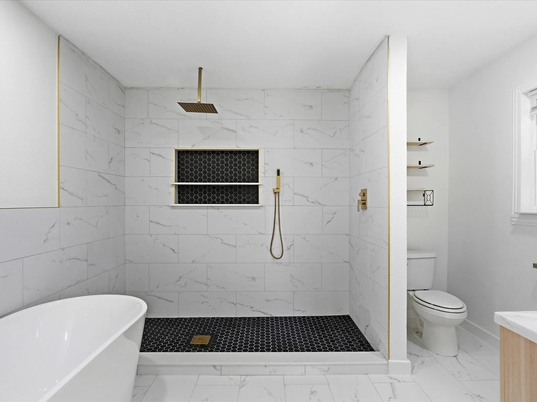 Walk-in shower featuring white marble-look tile walls, black hexagonal floor tile, and gold-finish fixtures