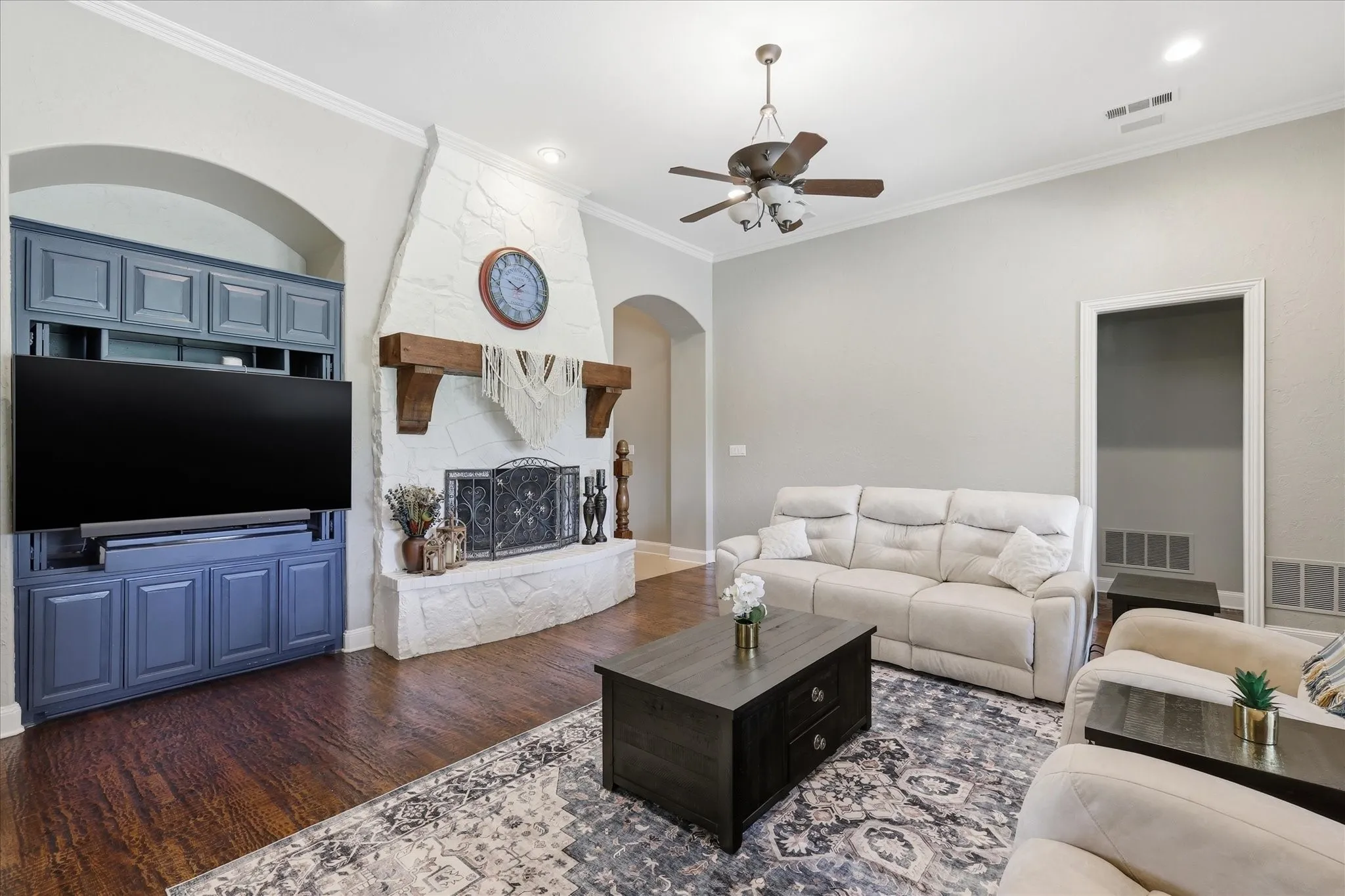 Living area featuring wood-finish flooring, a stone-clad fireplace with a substantial wood mantel, custom built-in cabinetry with an integrated media center, an arched doorway, and crown molding