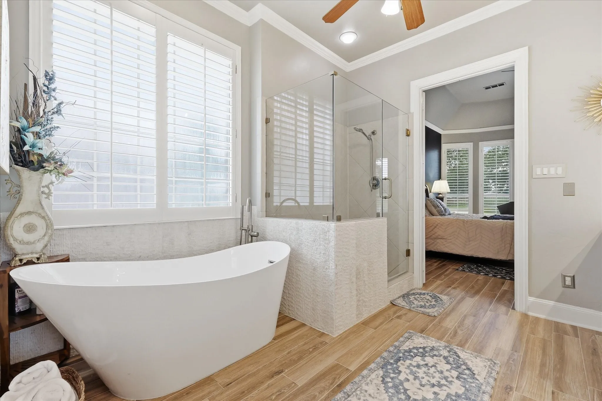 Contemporary freestanding soaking tub with brushed nickel fixtures, glass-enclosed shower with tiled surround, wood-finish flooring, and white plantation shutters