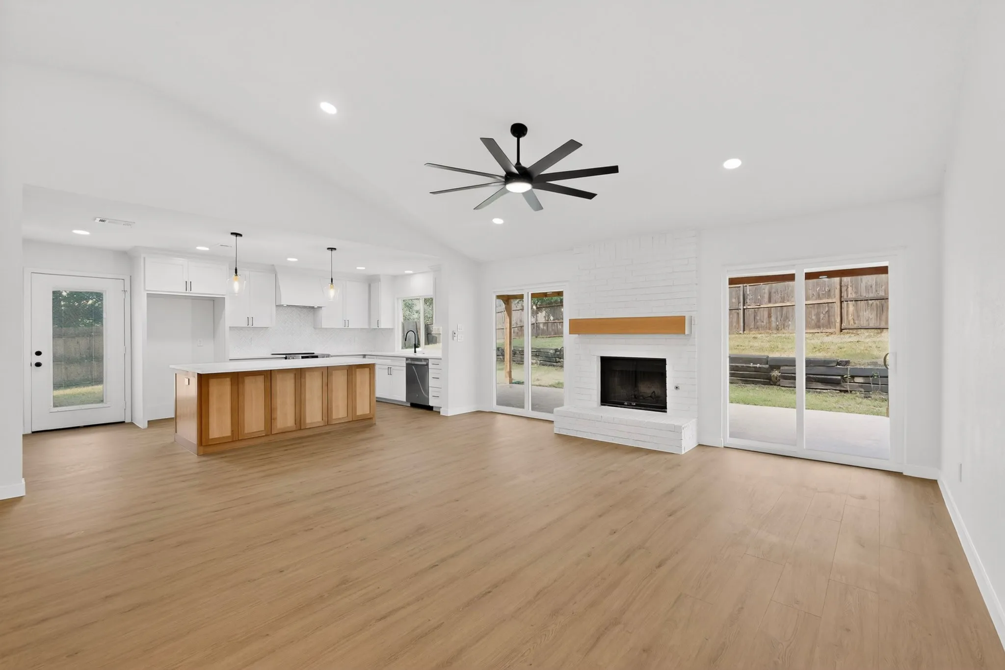 Expansive open-concept living area featuring wood-finish flooring, a white brick fireplace with a wood mantle, and sliding glass doors to the exterior