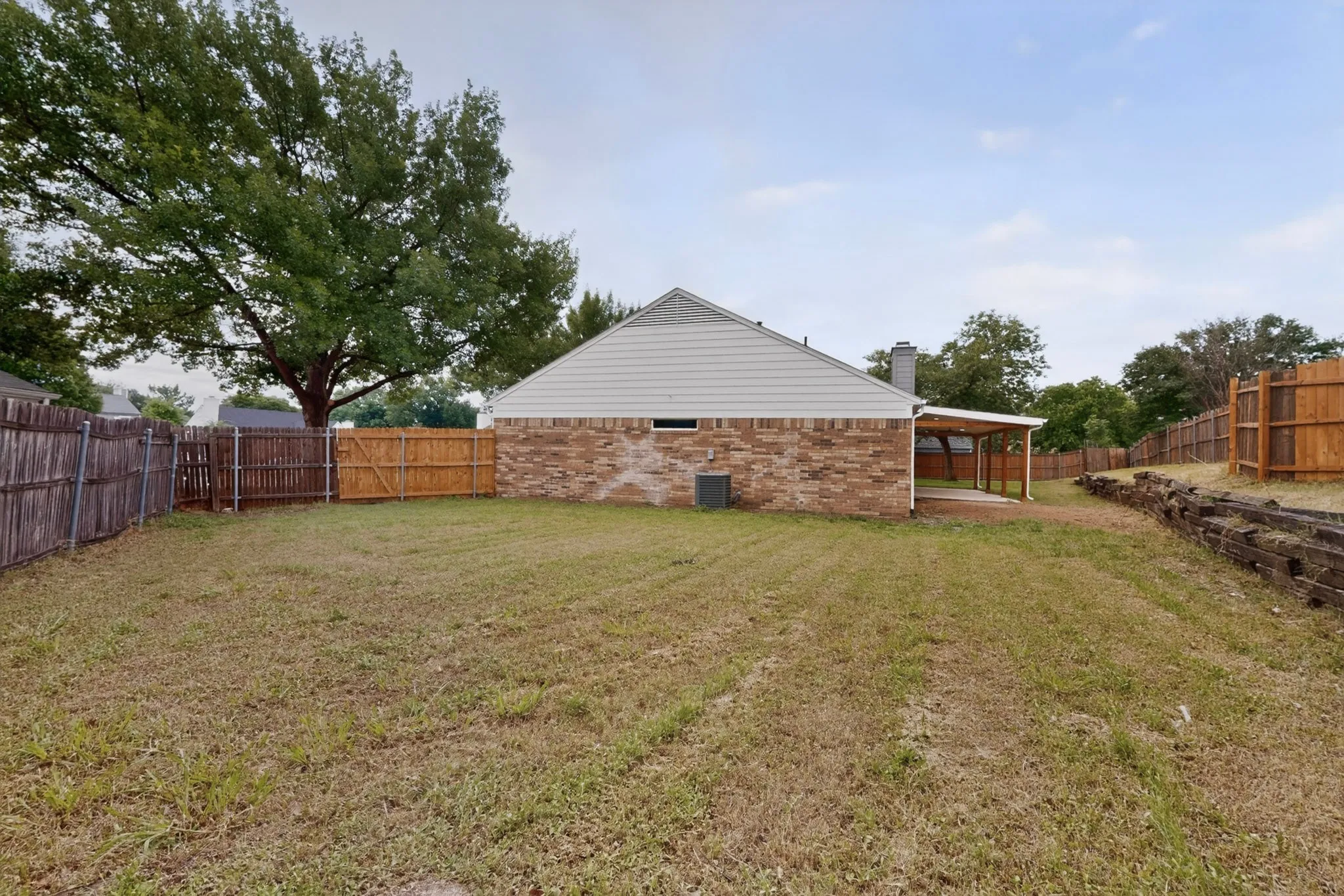 Expansive backyard featuring a partially covered patio, brick and siding exterior, a wood privacy fence, and mature perimeter trees